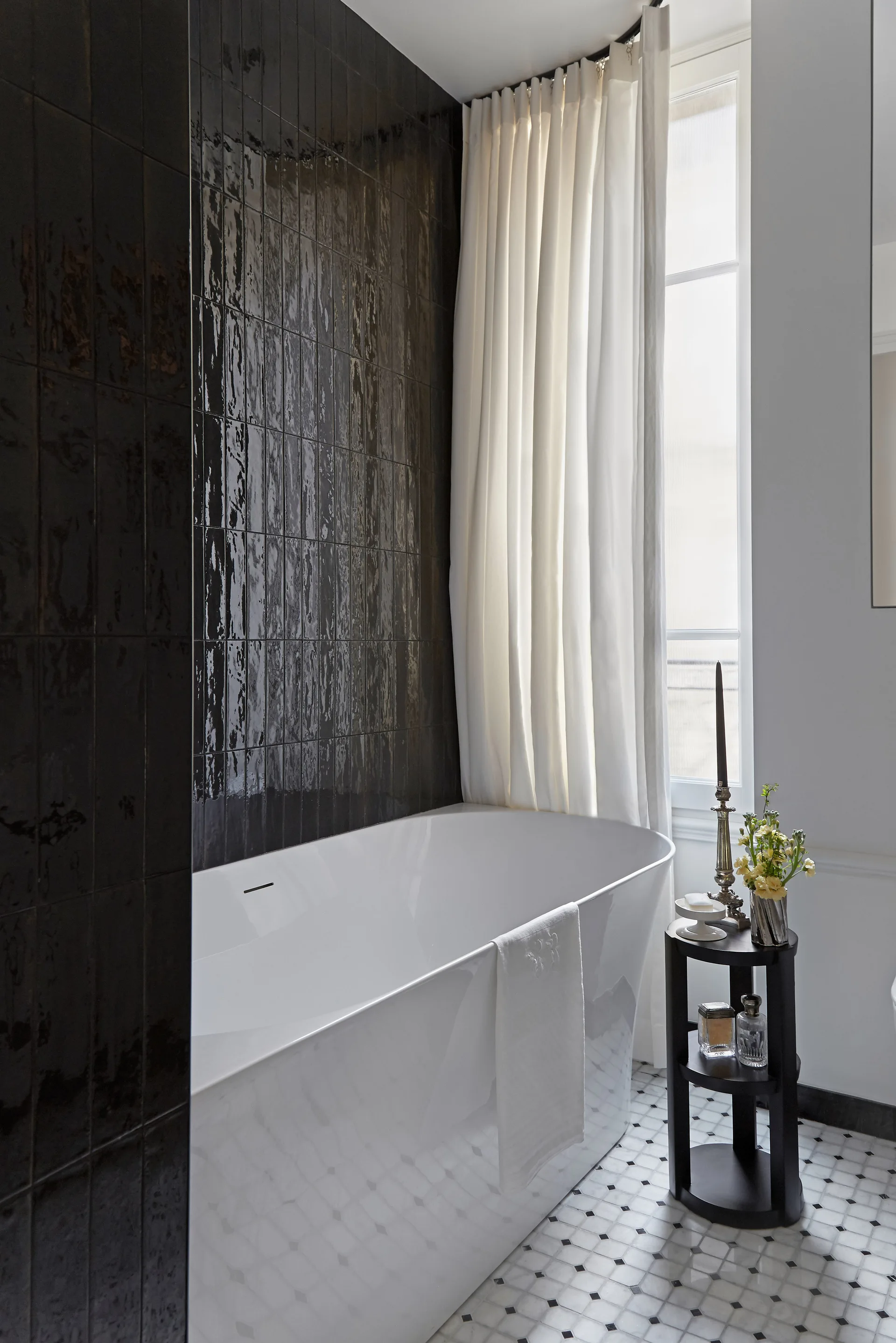 A white freestanding bathtub stands next to a high-gloss black tiled wall and a tall window with ivory curtains.