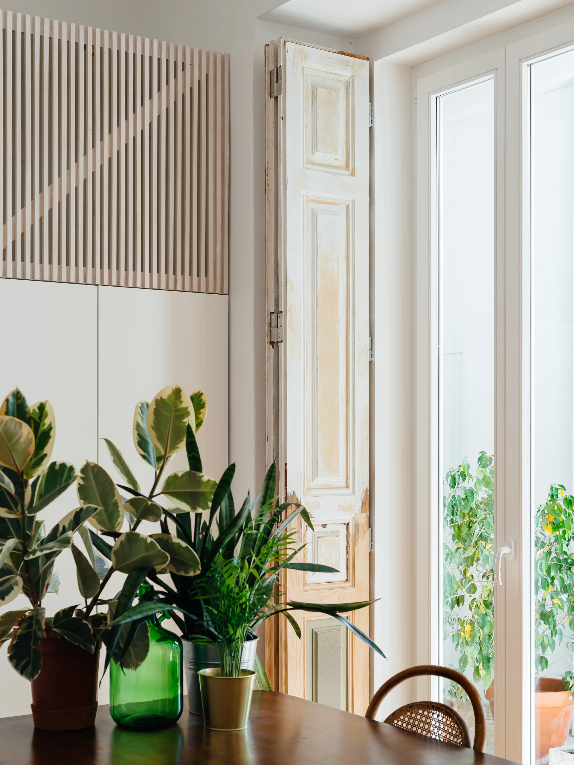 A dining area featuring a dark wood table with potted plants, a large window with a distressed wooden shutter, and vertical wood slat paneling on the wall.