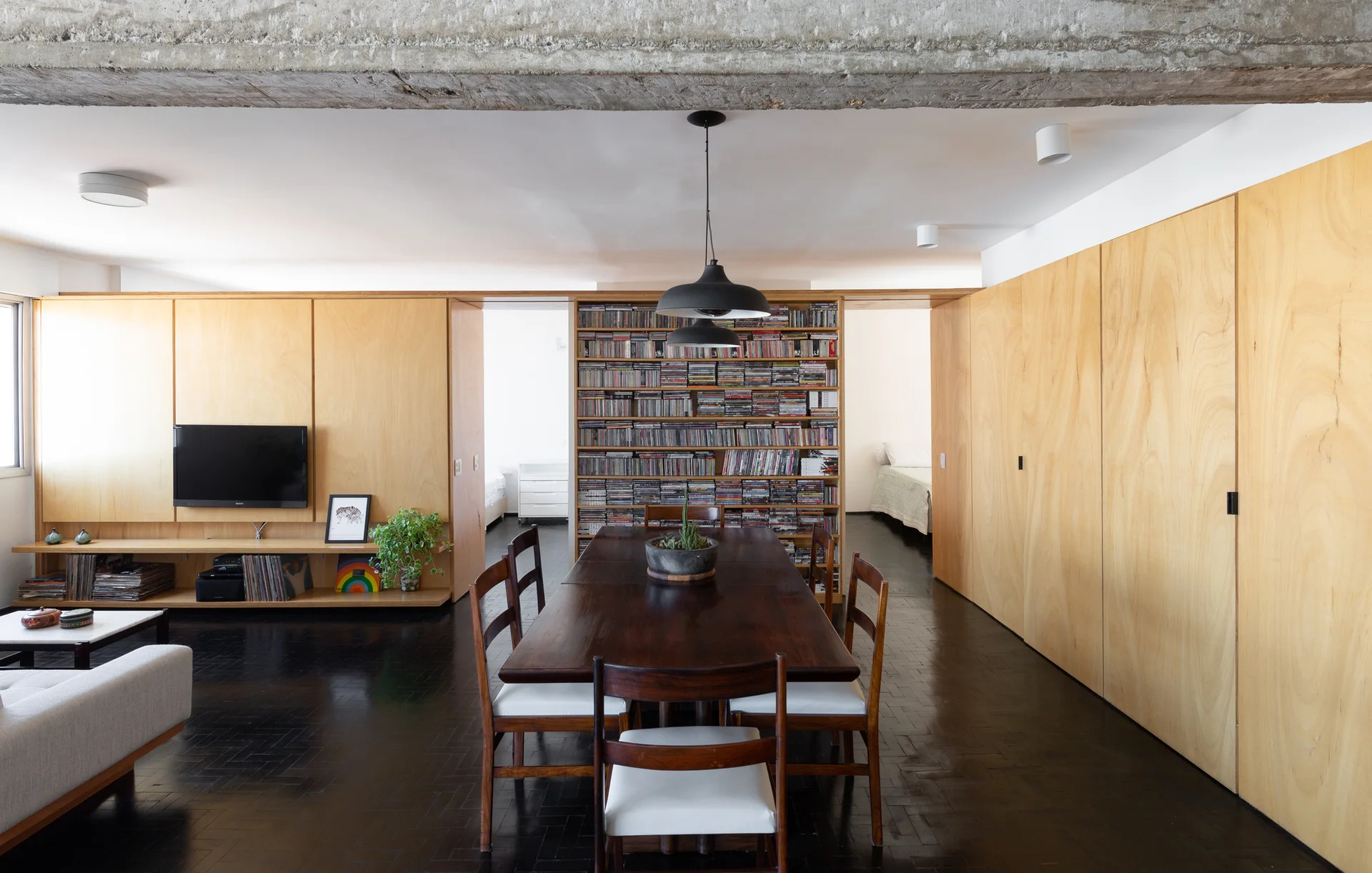 A modern dining room with a dark wood table, chairs, parquet flooring, light plywood paneled walls, and a rough exposed concrete ceiling beam.