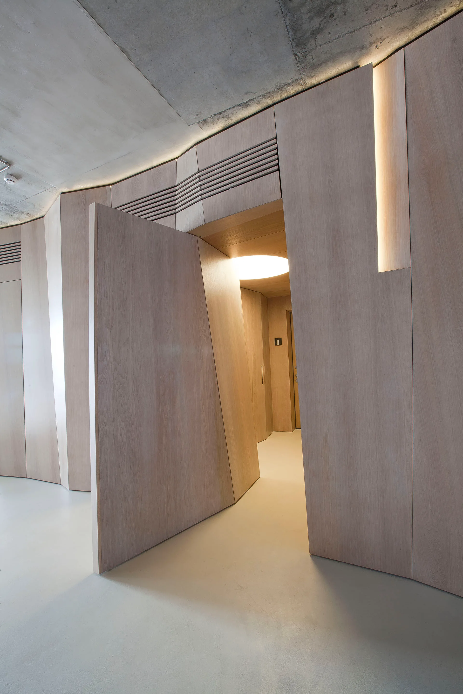 A minimalist hallway features custom light wood wall panels with an angular swinging door under an exposed concrete ceiling with integrated lighting.