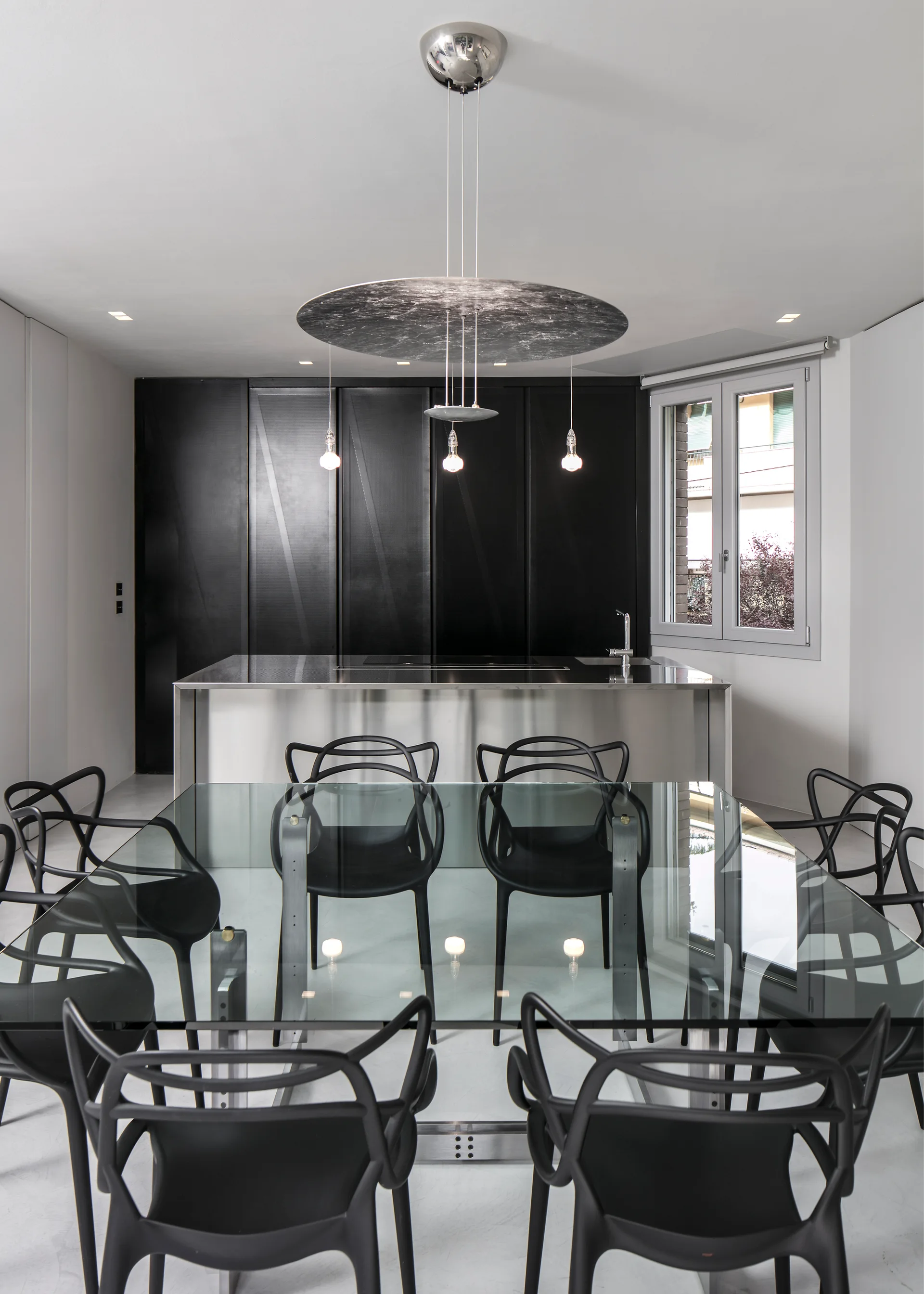 A modern dining room features a large glass-top table surrounded by black plastic chairs, set before a stainless steel kitchen island and dark cabinets.
