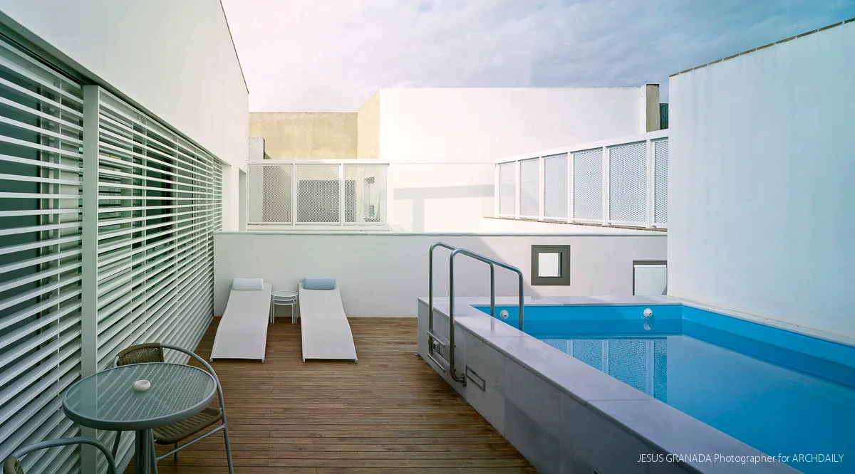 A modern rooftop terrace featuring a raised blue swimming pool, wooden plank decking, white sun loungers, and white louvered walls.