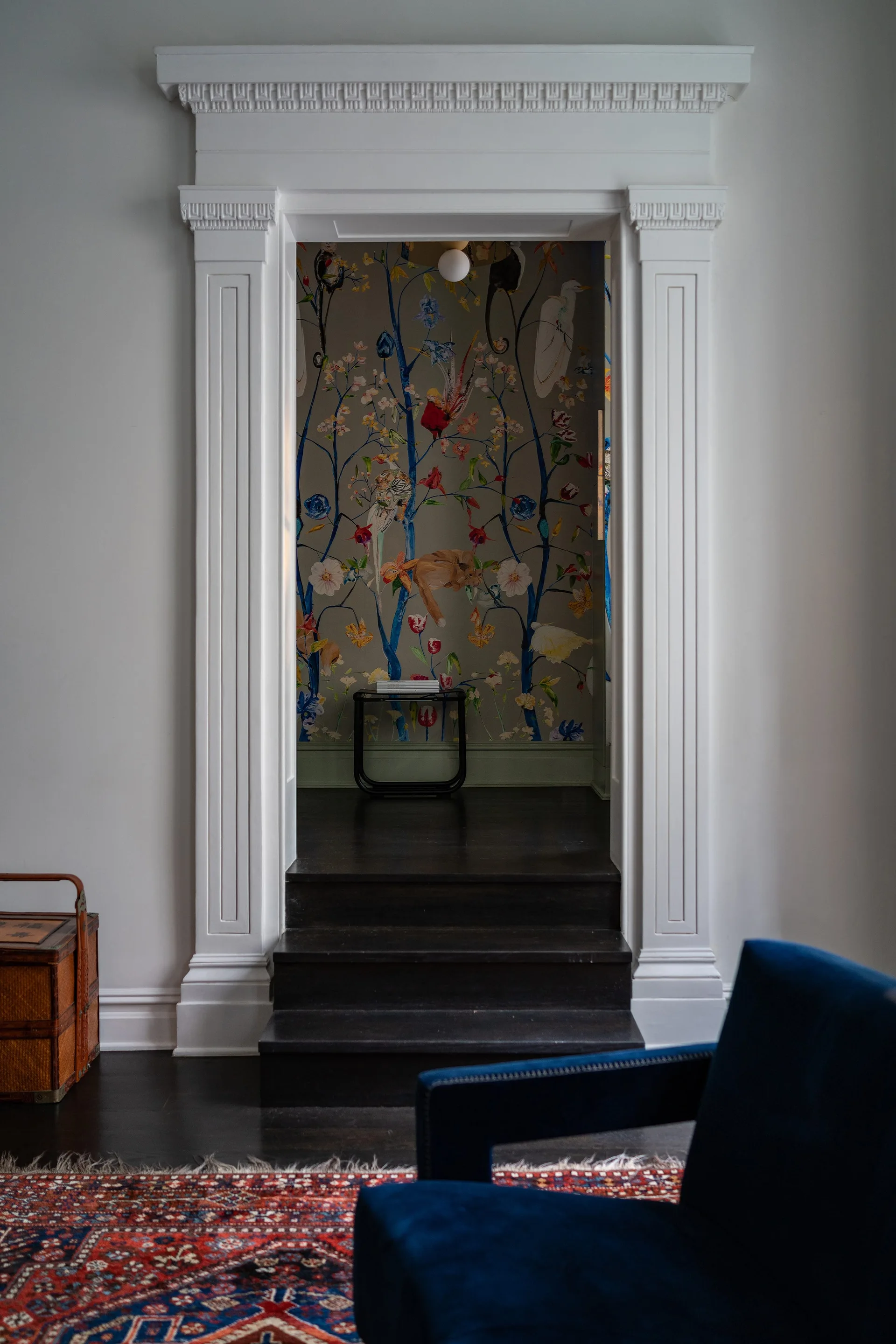 A classical white cased opening framed by dark wood steps leads to an entryway wrapped in botanical wallpaper featuring birds and trees.