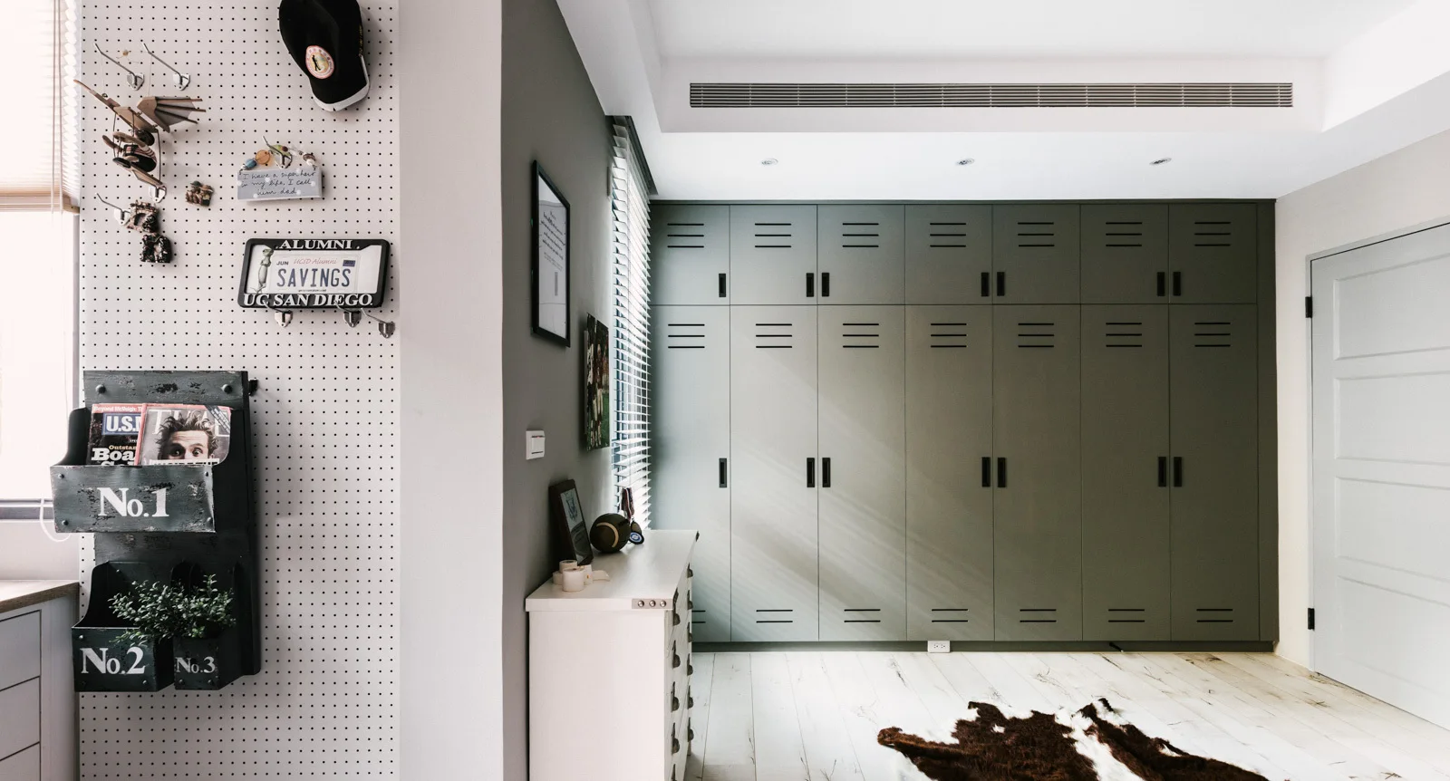 A bedroom features an olive green locker storage wall, a white pegboard organizer, and light wood floors with a faux cowhide rug.