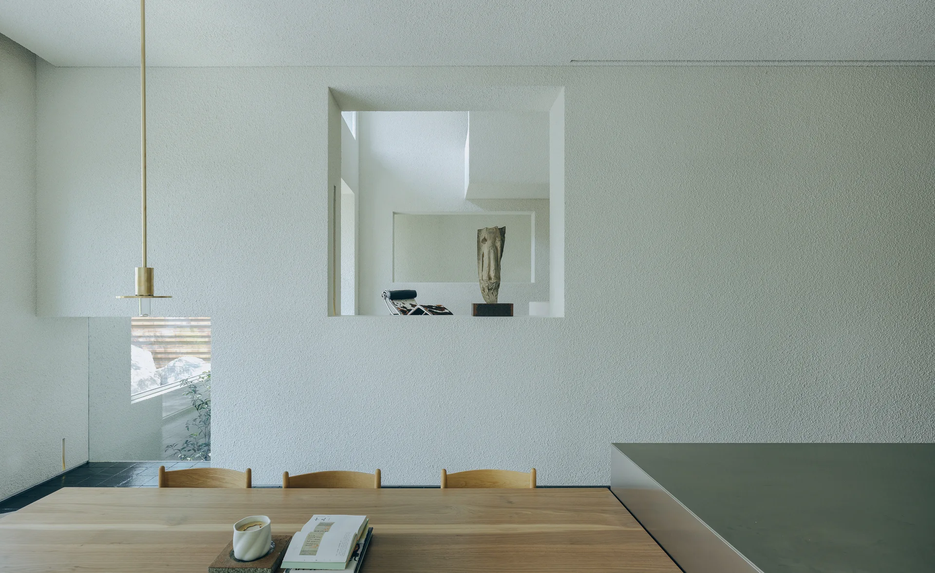 A light wood dining table rests before a stippled off-white plaster wall featuring a square aperture that reveals a sculpture in the room beyond.