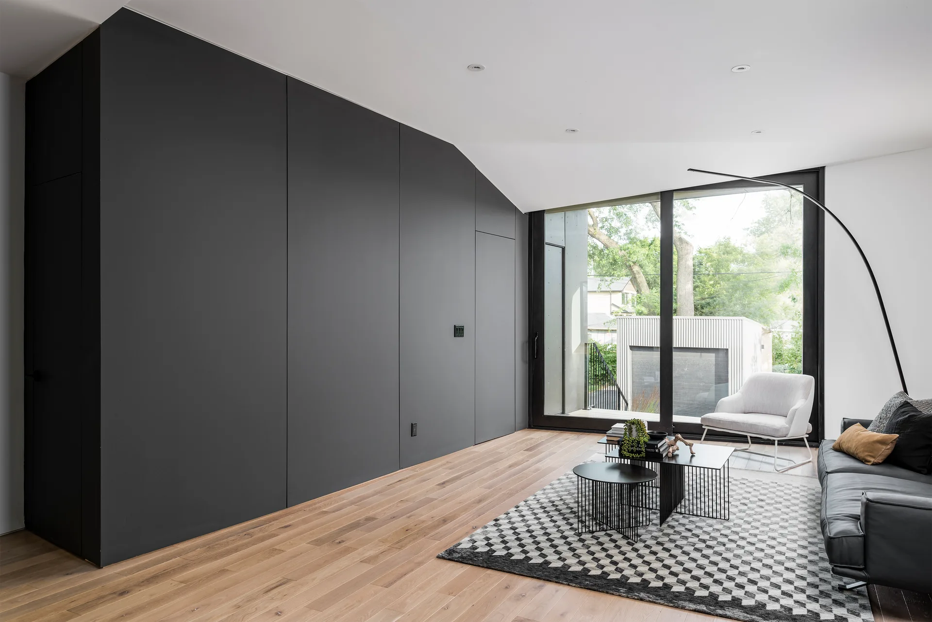 A modern living room featuring a matte charcoal storage wall, a checkered area rug, a black leather sofa, and a large glass window.