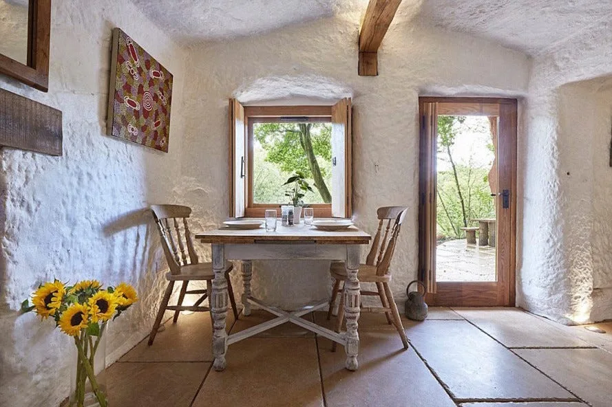 A rustic dining area features a wooden table and two chairs set against highly textured white plaster walls next to a shuttered window.