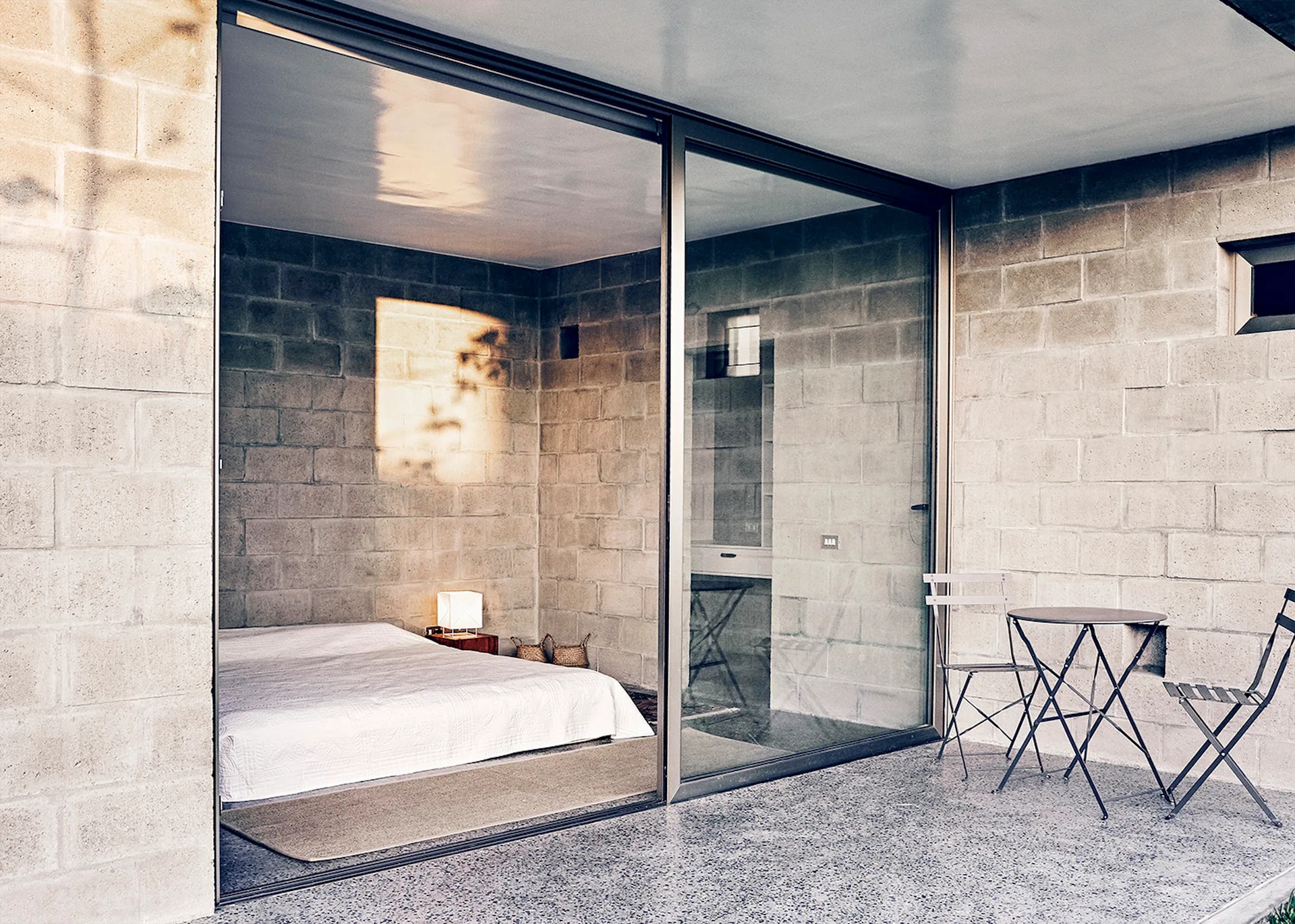A minimalist bedroom with concrete block walls and a low bed connects to an outdoor concrete patio with a black metal bistro set.
