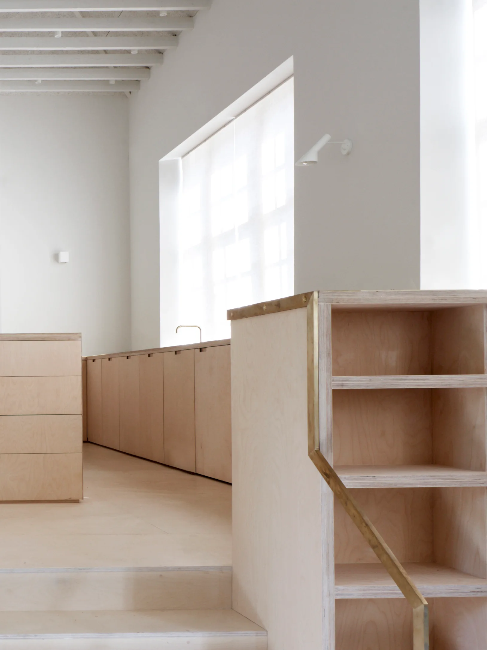 Minimalist kitchen featuring light plywood cabinetry, open shelving accented with a thin brass trim, and white walls under exposed ceiling beams.