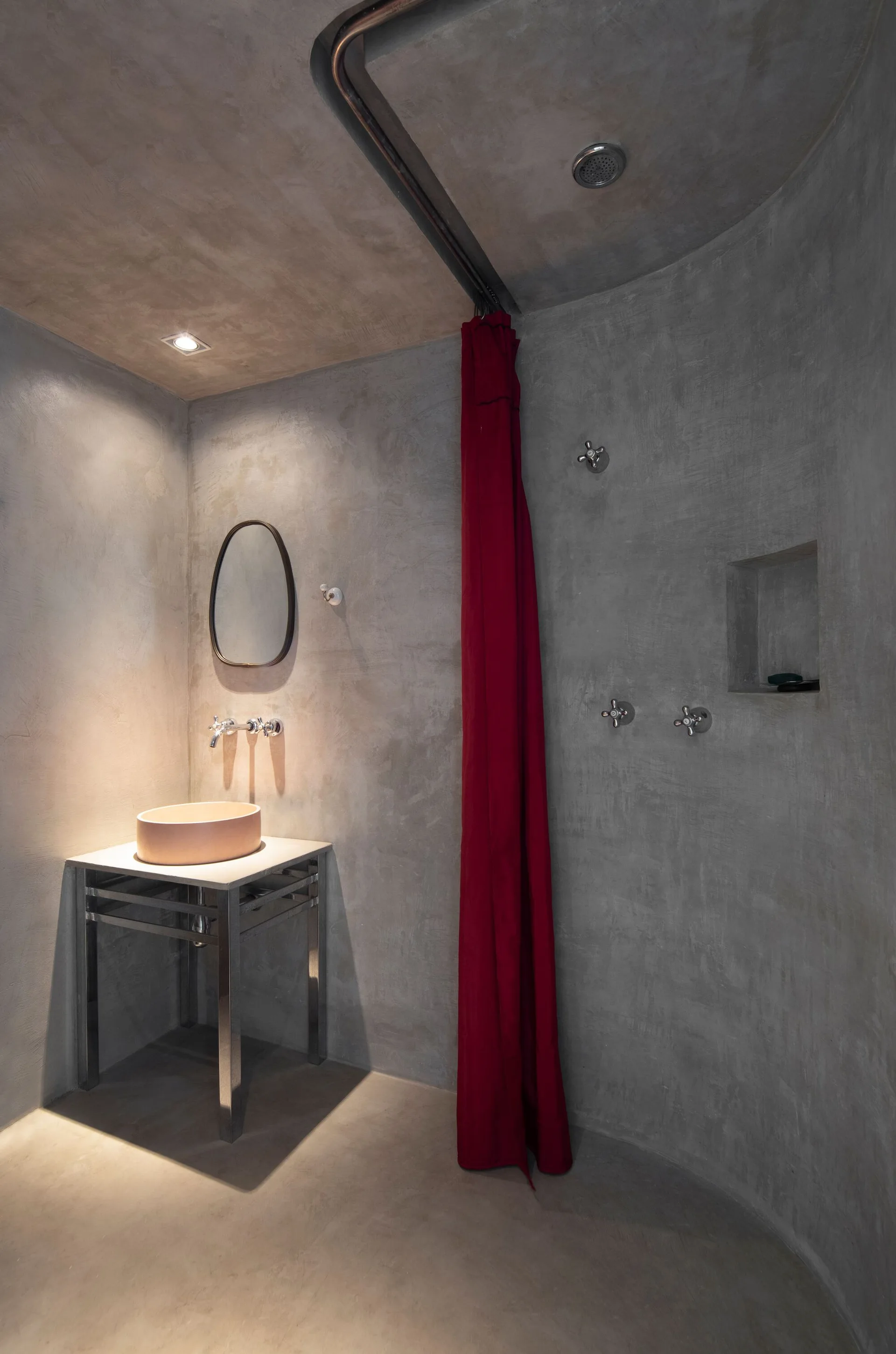 A minimalist grey concrete bathroom features a metal vanity with a round peach vessel sink, an oval mirror, and a long red shower curtain.