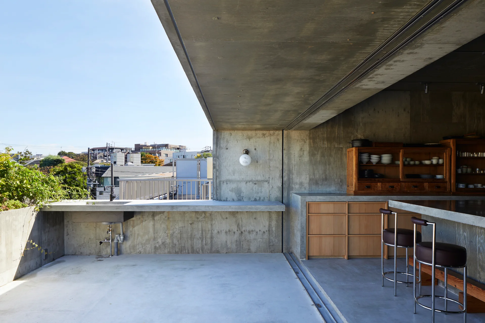 An open-air concrete patio features an outdoor kitchen with wooden cabinetry, a counter sink, and chrome bar stools under a heavy concrete overhang.