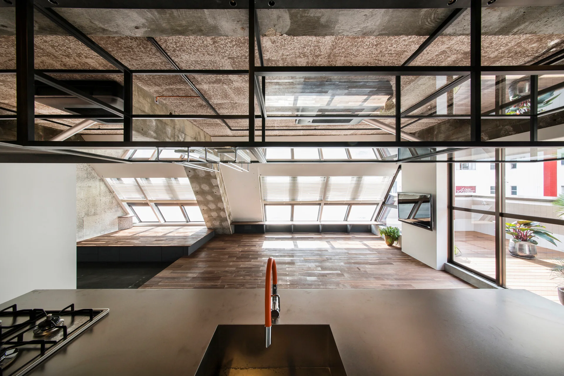 An industrial loft kitchen features a dark island with an orange faucet, under an exposed concrete ceiling and angled skylight windows.