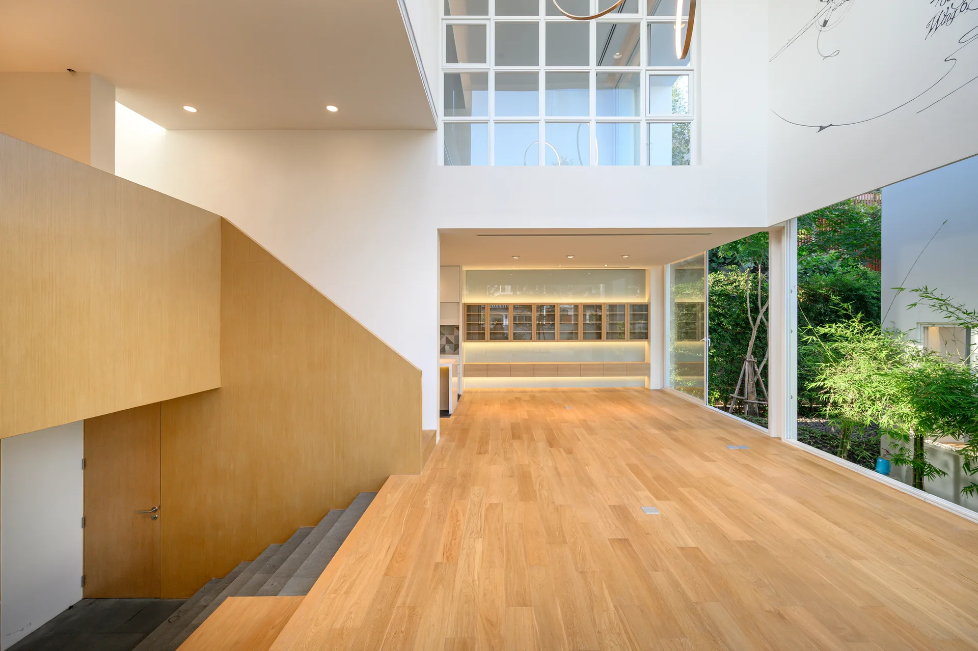 A modern open-plan living room features light wood floors, a wood-paneled staircase wall, and large glass windows overlooking an outdoor garden.