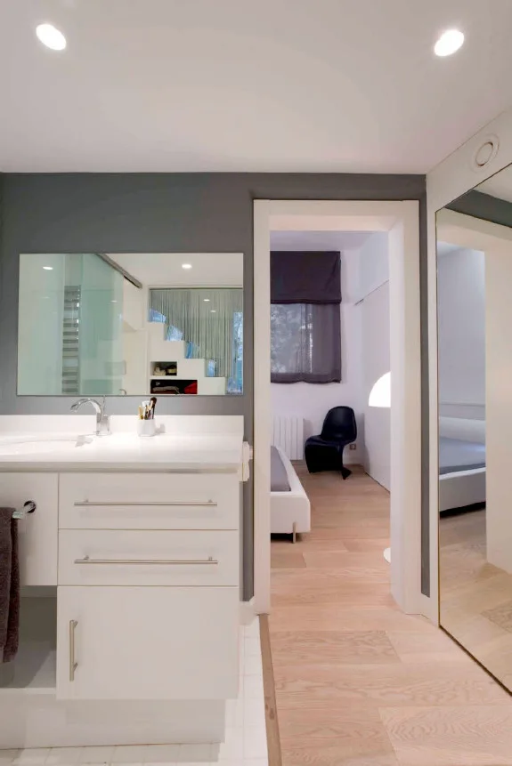 A white bathroom vanity with a chrome faucet stands against a grey wall next to a doorway looking into a bedroom with light wood floors.