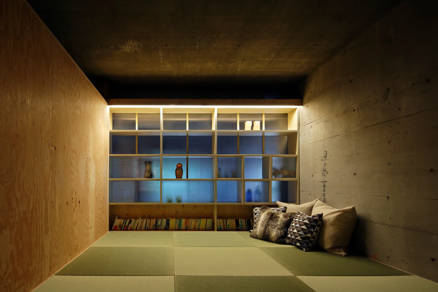 A minimalist lounge room features woven green tatami mats, a light wood plywood wall, an exposed concrete wall, and a backlit wooden grid shelf unit.