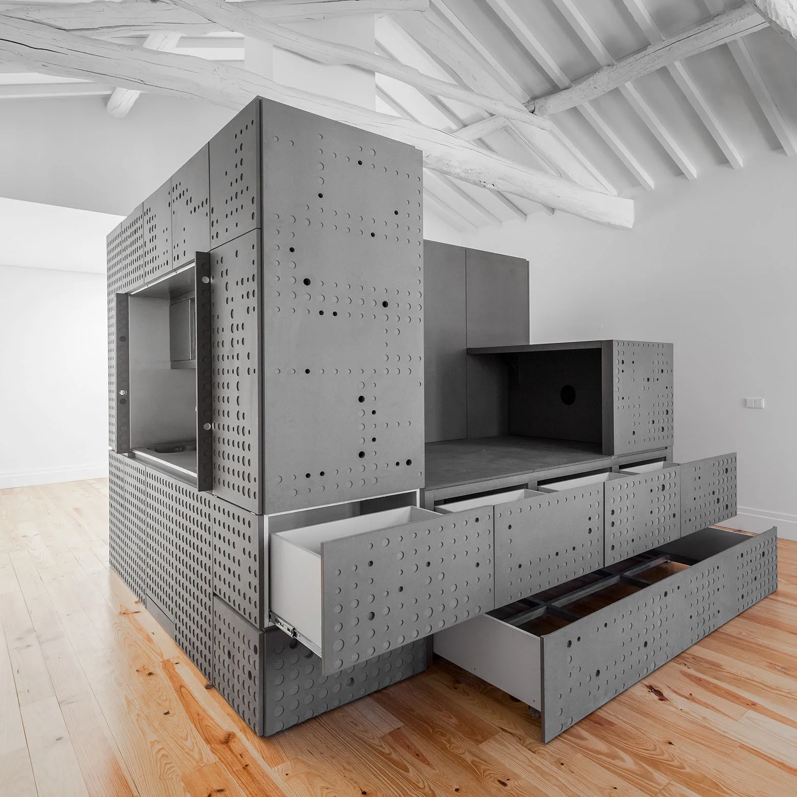 A large grey modular cabinet unit with perforated circular patterns and open drawers stands on a light wood floor beneath white exposed ceiling beams.