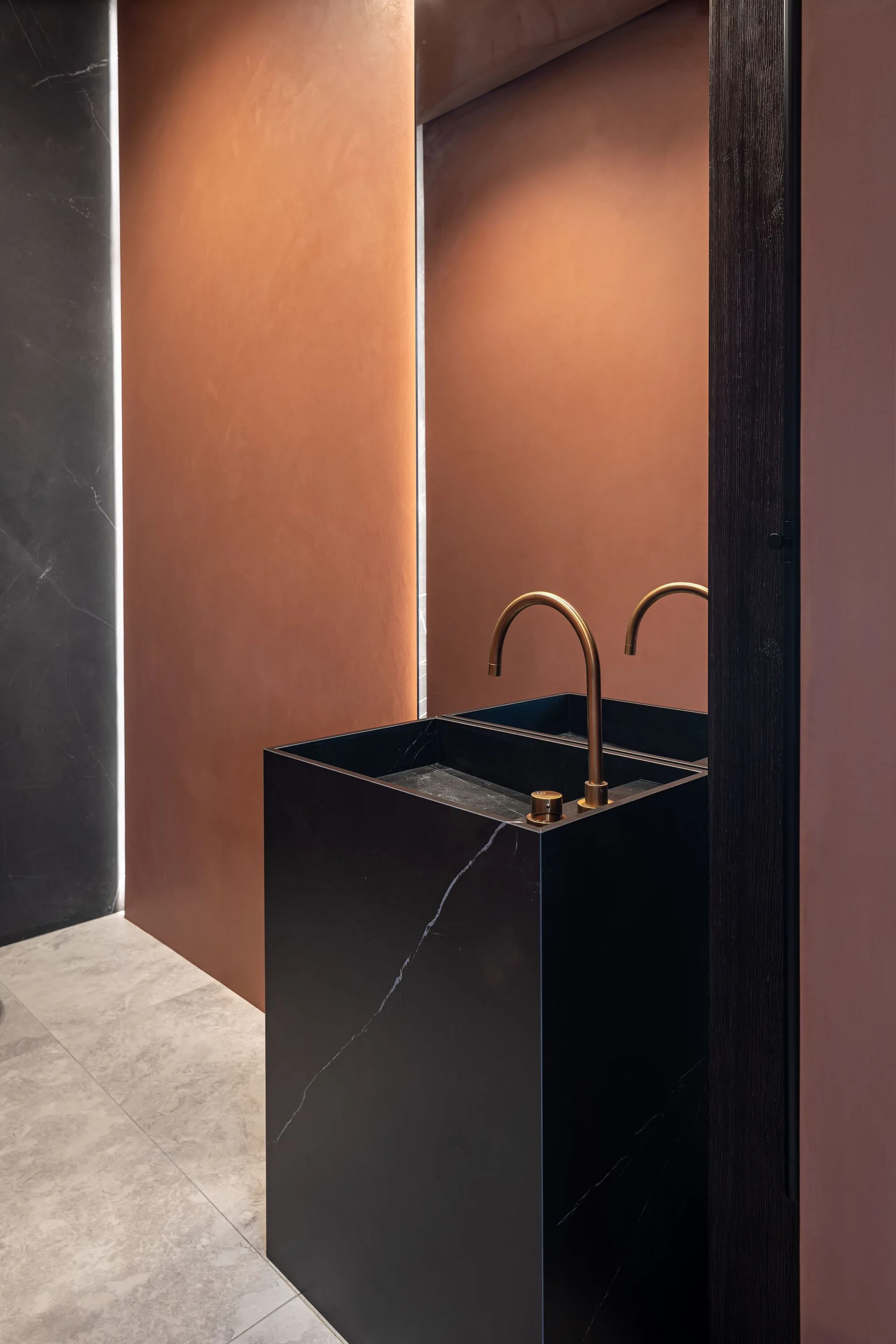 A minimalist powder room features a black marble pedestal sink with a gold faucet against a smooth copper wall panel and a large vertical mirror.