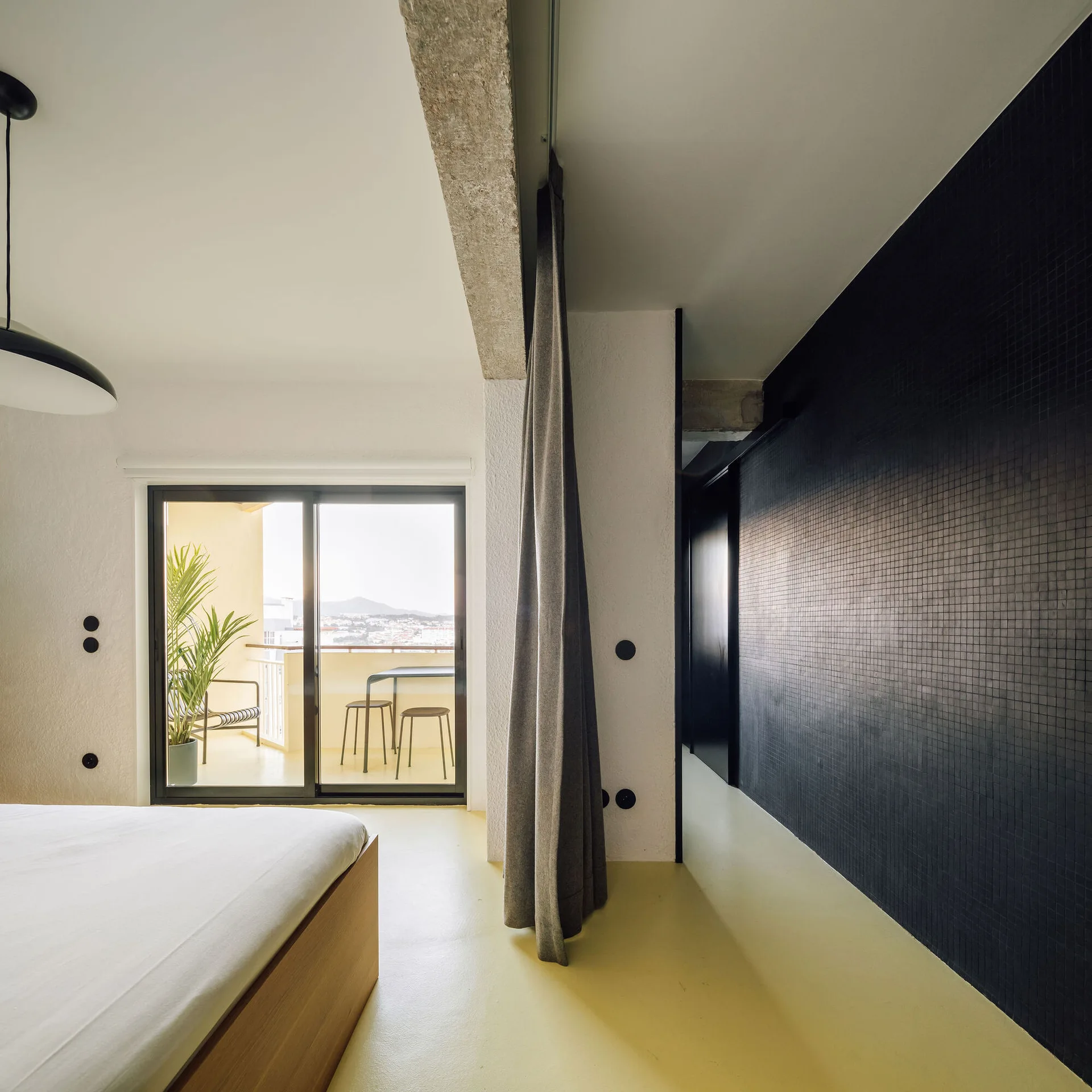 A minimalist bedroom featuring a bright yellow floor, a black grid-tiled accent wall, and a sliding glass door that leads to a balcony.