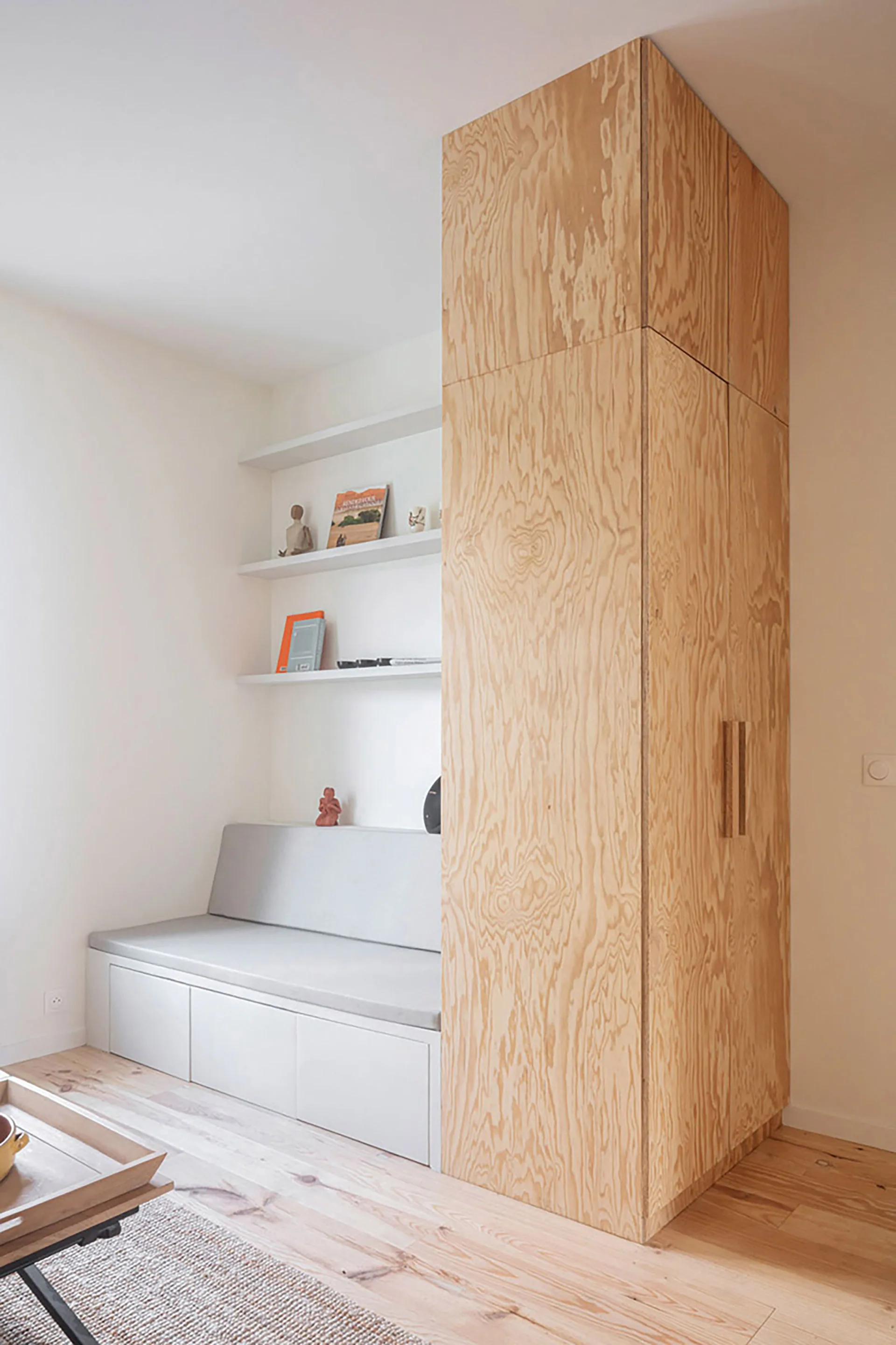 A tall plywood storage wardrobe stands next to a white built-in bench seat with gray cushions and floating shelves on a white wall.