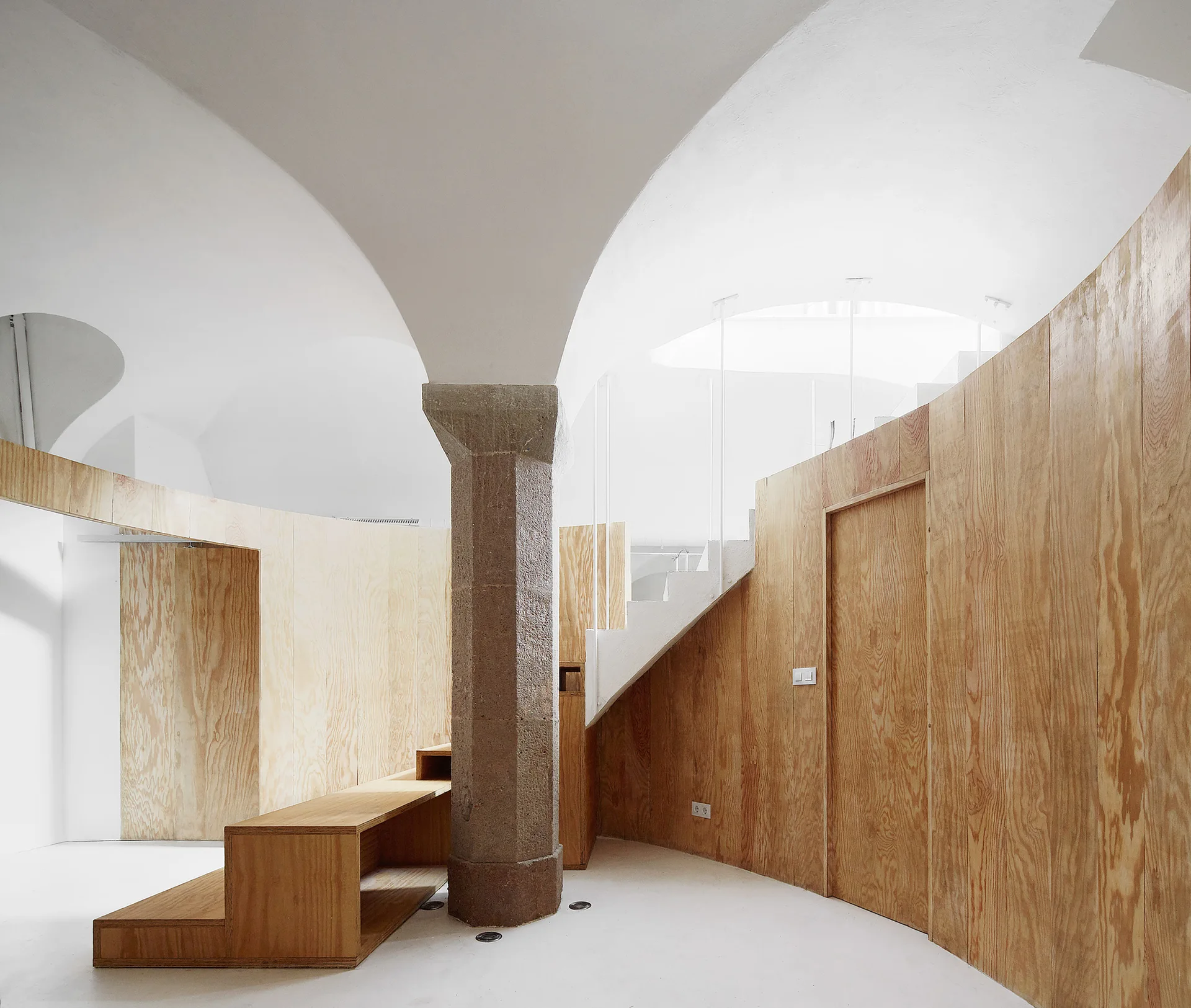 A minimalist room features curved plywood wall paneling, a central stone column under a white vaulted ceiling, and an integrated white staircase.