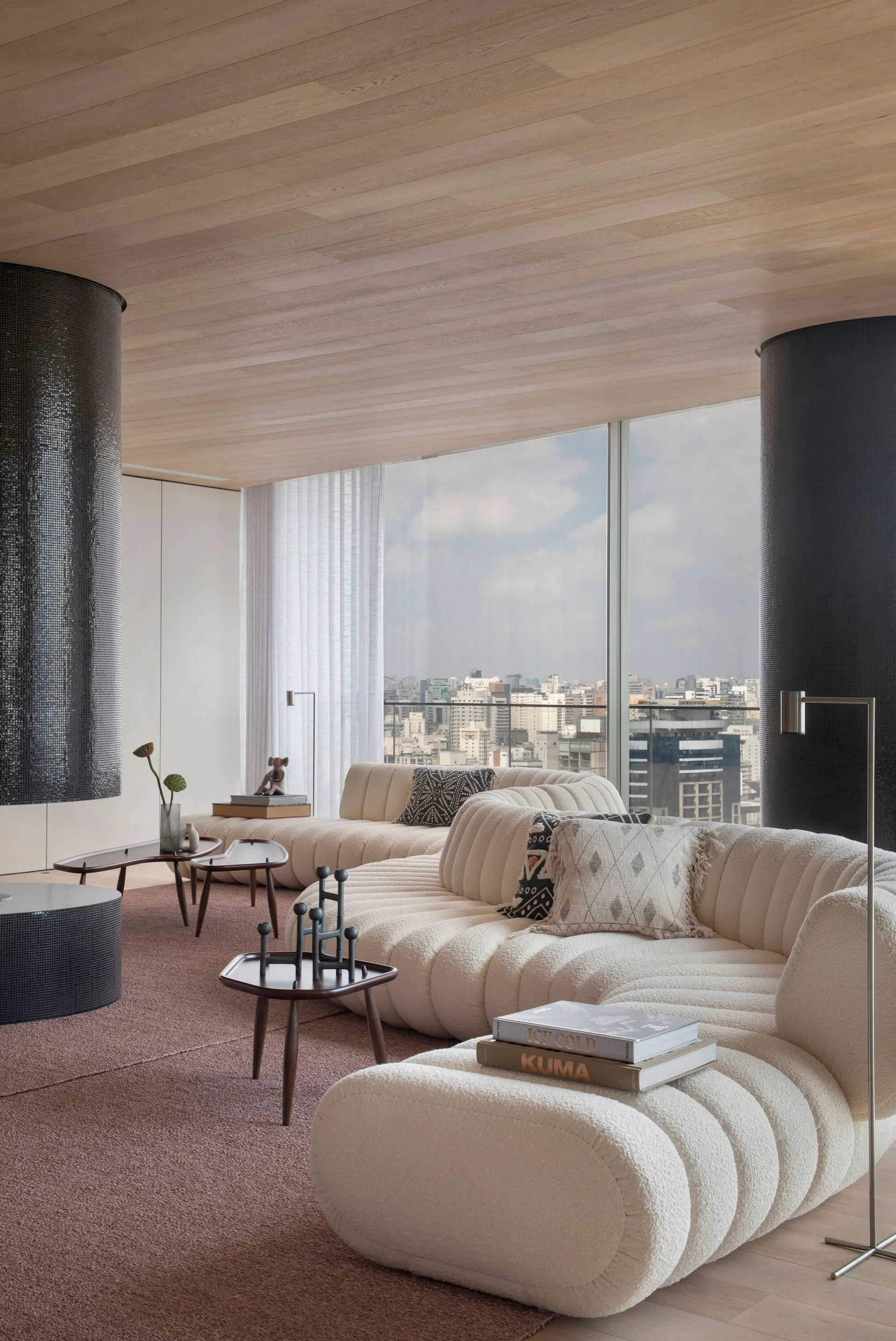 A spacious living room featuring a curved cream sectional sofa, low wood tables, black tiled columns, and floor-to-ceiling windows with a city view.