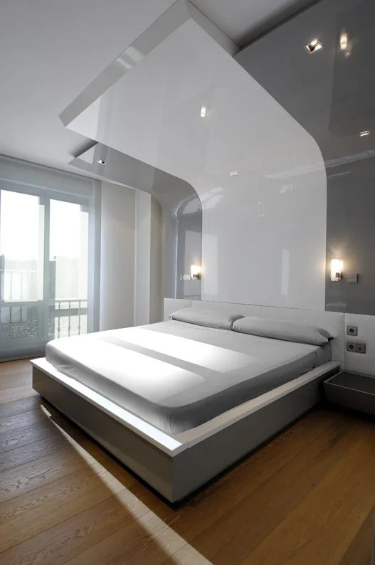 A minimalist bedroom with a low grey platform bed framed by a glossy, curved white and grey architectural canopy over wide plank wood floors.