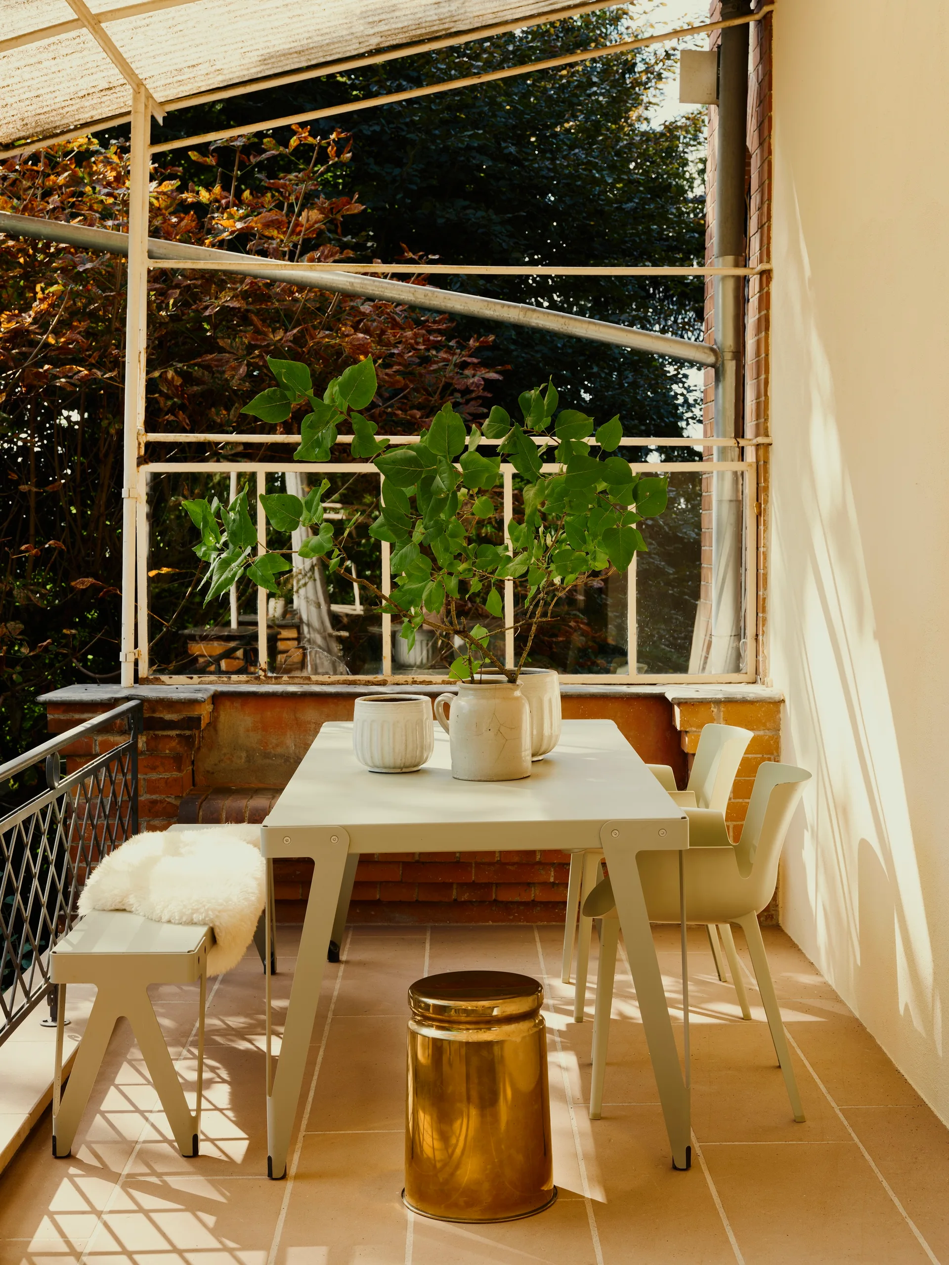 A covered outdoor patio features a sage green metal dining table and chairs, a bench with a faux fur throw, and a gold metallic stool.