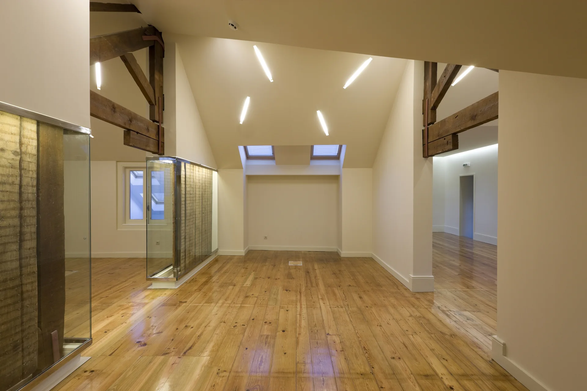 An empty attic room featuring polished wood flooring, exposed dark timber trusses, white sloped walls, skylights, and a modern glass partition.