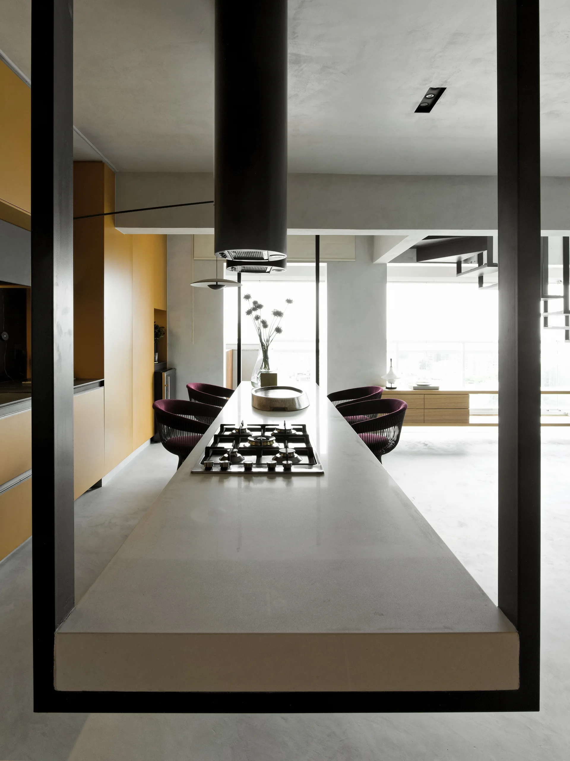 A modern grey kitchen featuring a long concrete island with an integrated cooktop, black cylindrical range hood, and tall yellow cabinets.