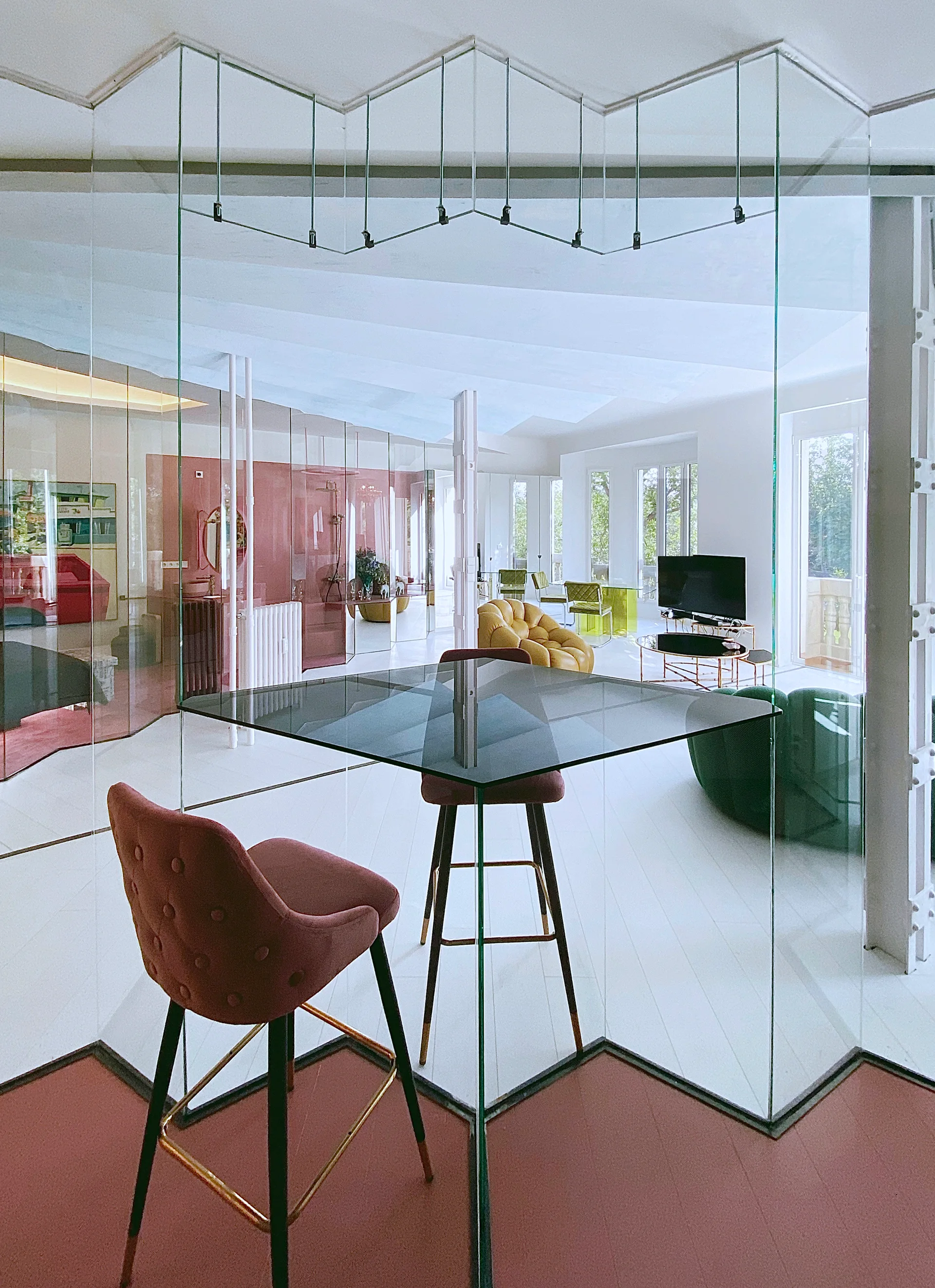 Modern open-plan living room featuring clear glass partition walls, a square black glass table with a dusty pink tufted bar stool, and white floors.
