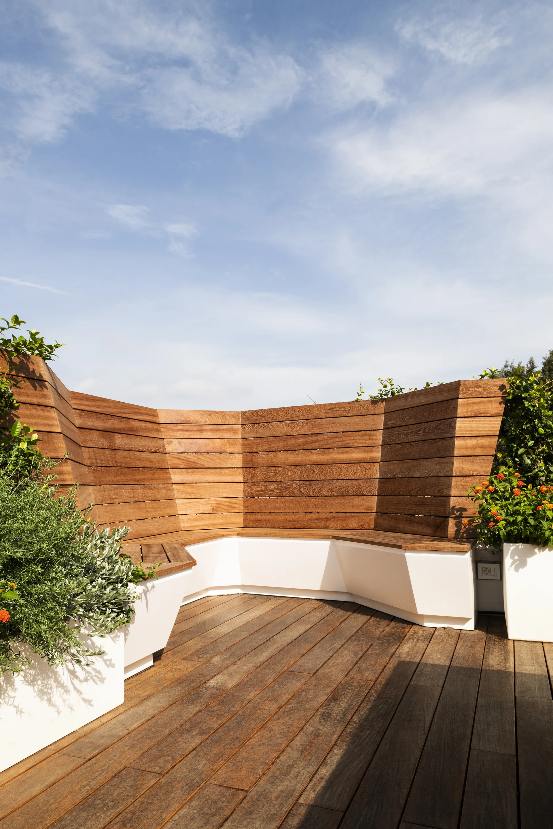 A curved built-in wooden bench with integrated white planters stands on a wood-plank deck under a blue sky.