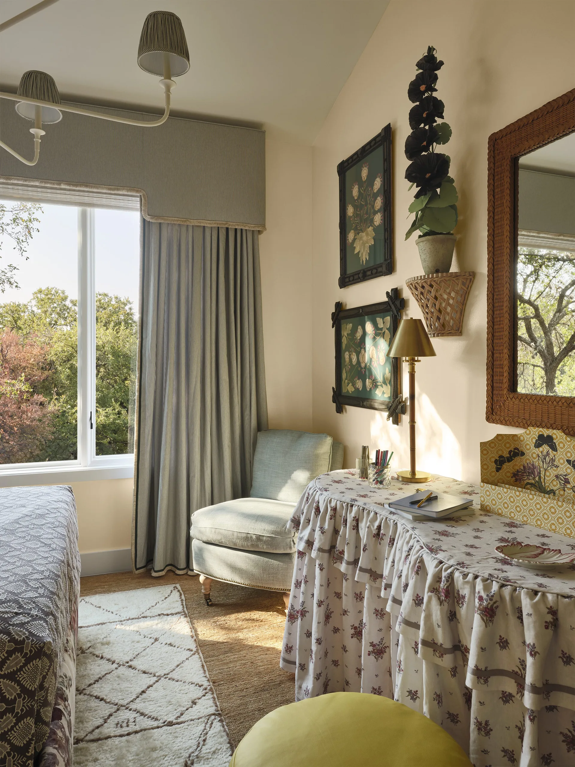 A sunlit bedroom corner features a floral skirted vanity table, a soft blue armchair, striped curtains, and botanical art prints on cream walls.
