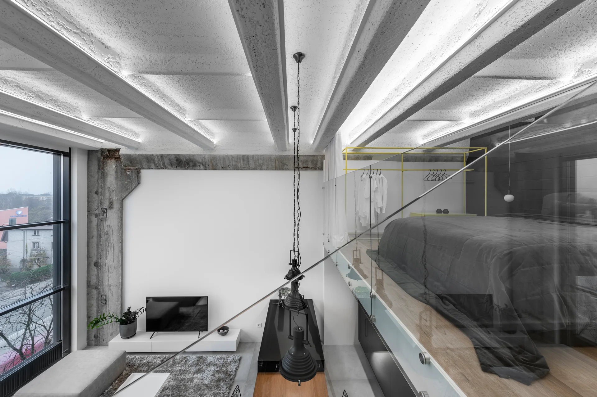 A multi-level industrial loft features an elevated bedroom with a glass partition overlooking a lower-level living area with exposed concrete beams.