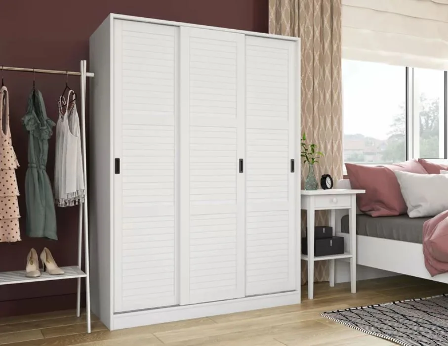 A white wardrobe with three slatted sliding doors stands next to a garment rack and nightstand against a dark burgundy wall.
