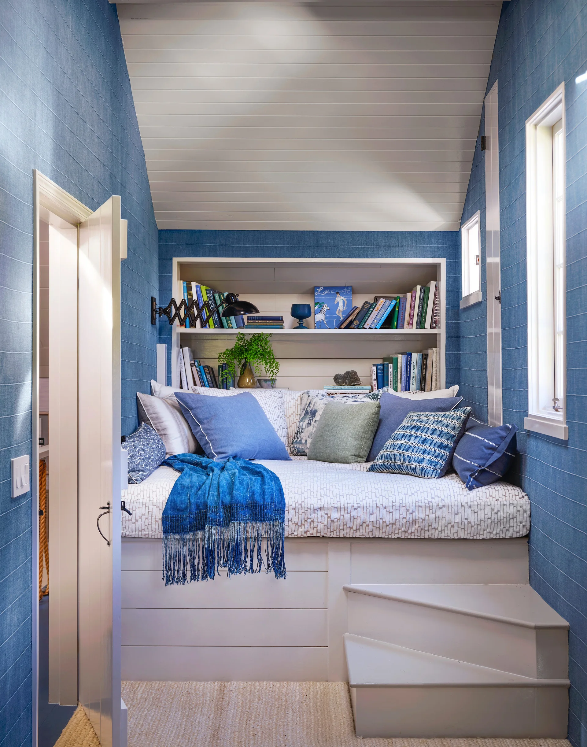 A built-in daybed nook features blue textured wallpaper, white recessed bookshelves, steps leading up, and a collection of blue and patterned pillows.