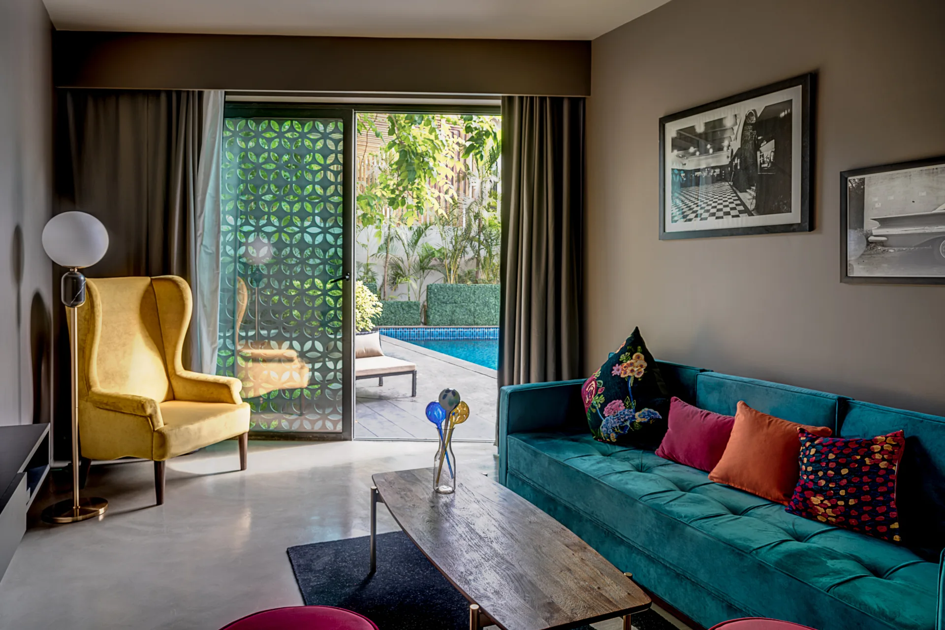 A living room features a teal velvet sofa, a yellow wingback chair, and sliding glass doors that open to an outdoor swimming pool.