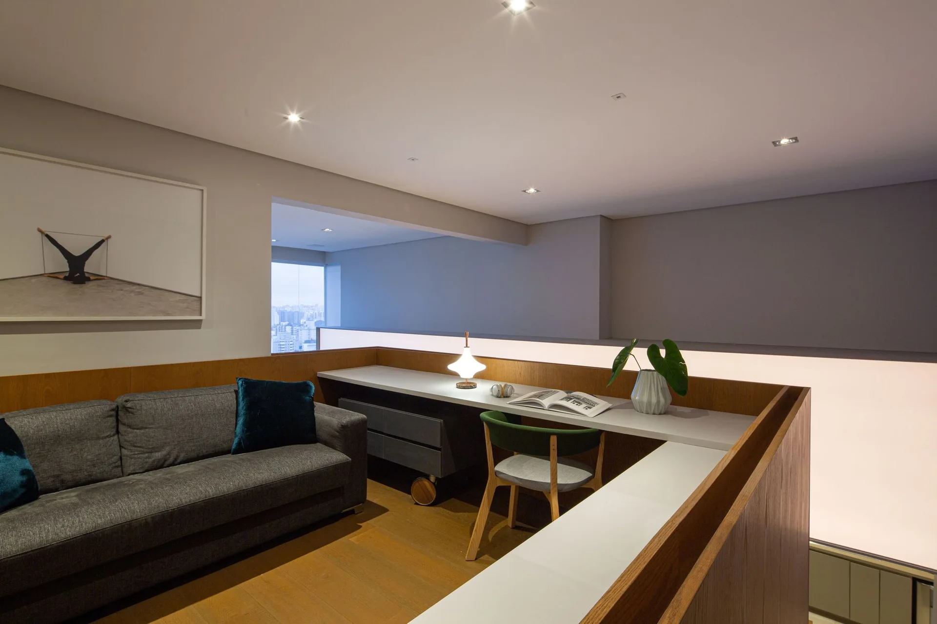 A modern loft home office featuring a grey fabric sofa, wood wall paneling, and a long built-in desk with a green chair.