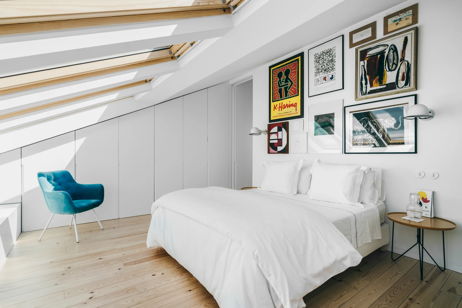 Bright attic bedroom with a white bed, sloped wood-framed skylights, seamless white closets, and a colorful gallery art wall.
