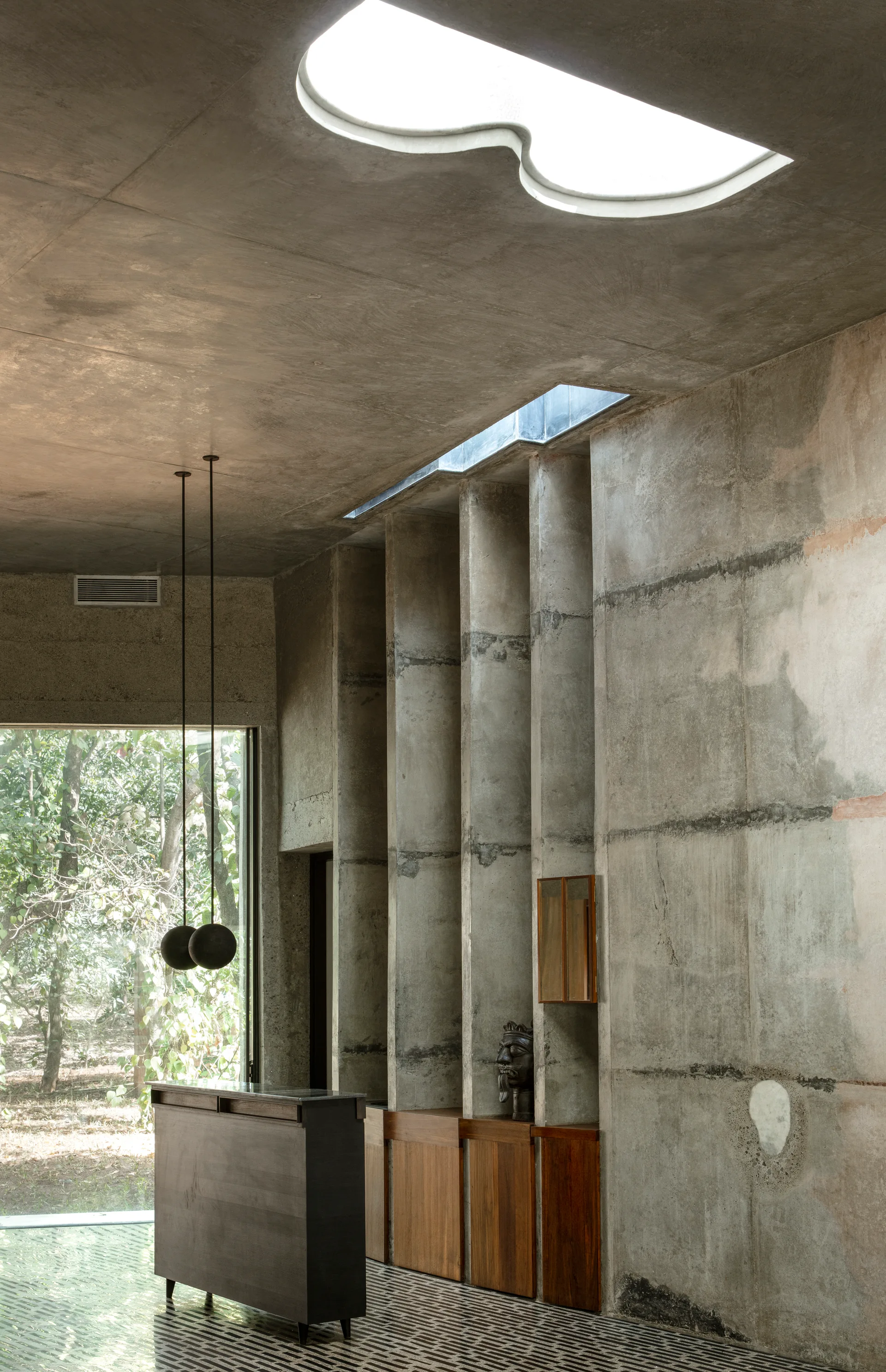 An interior room constructed of raw concrete features a curved ceiling skylight, vertical wall pillars, and integrated wooden cabinets.