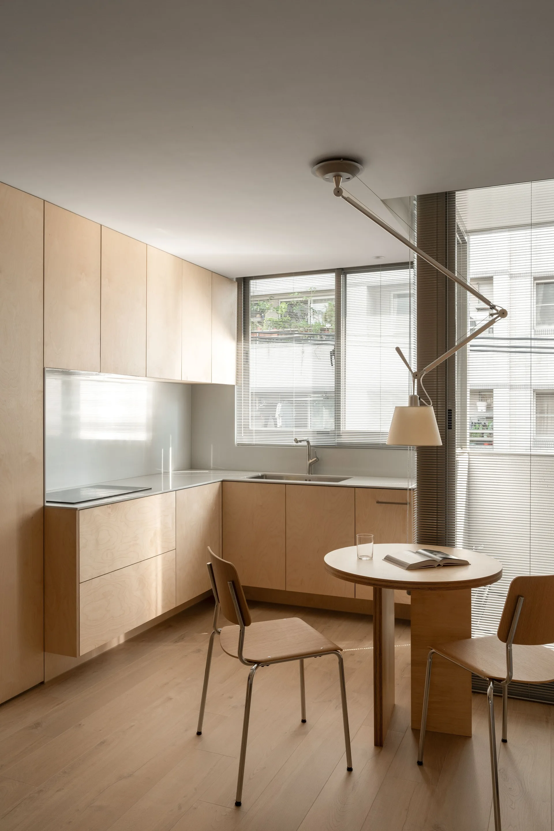 A minimalist kitchen with light-toned plywood cabinets, a round wooden dining table with two chairs, and an adjustable pendant light.