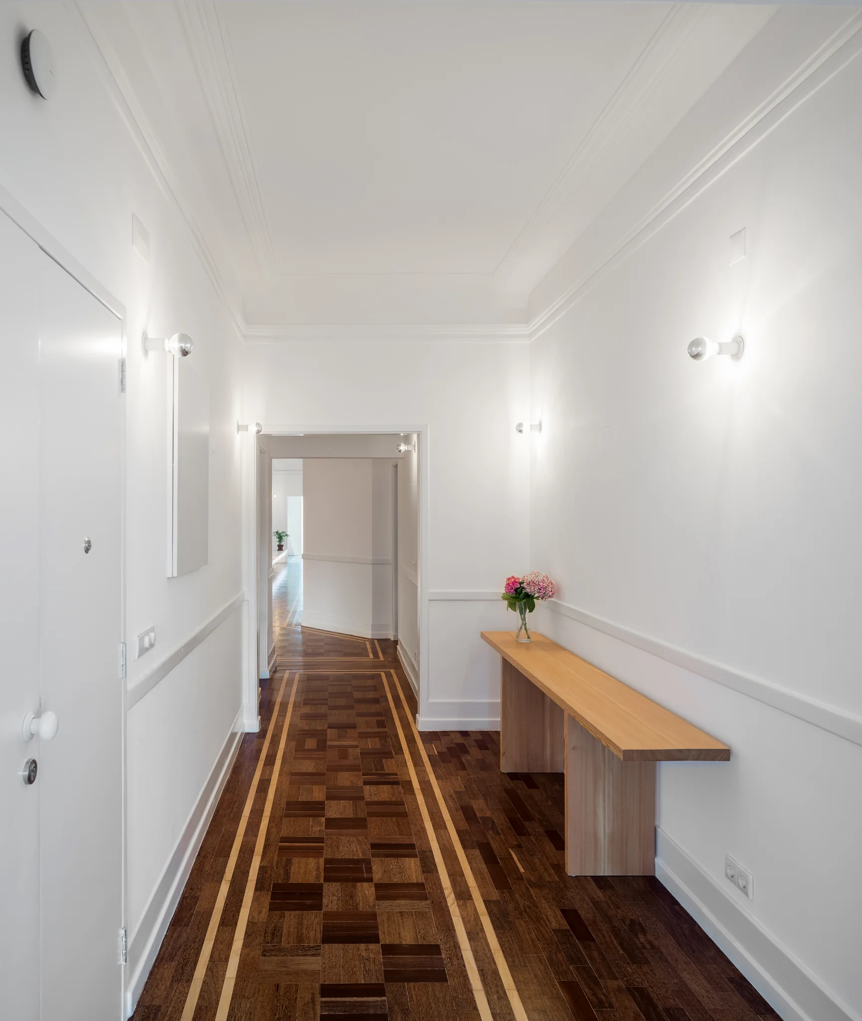 A long white residential corridor features a minimalist wood console table, simple wall sconces, and dark patterned wood flooring with light-toned borders.