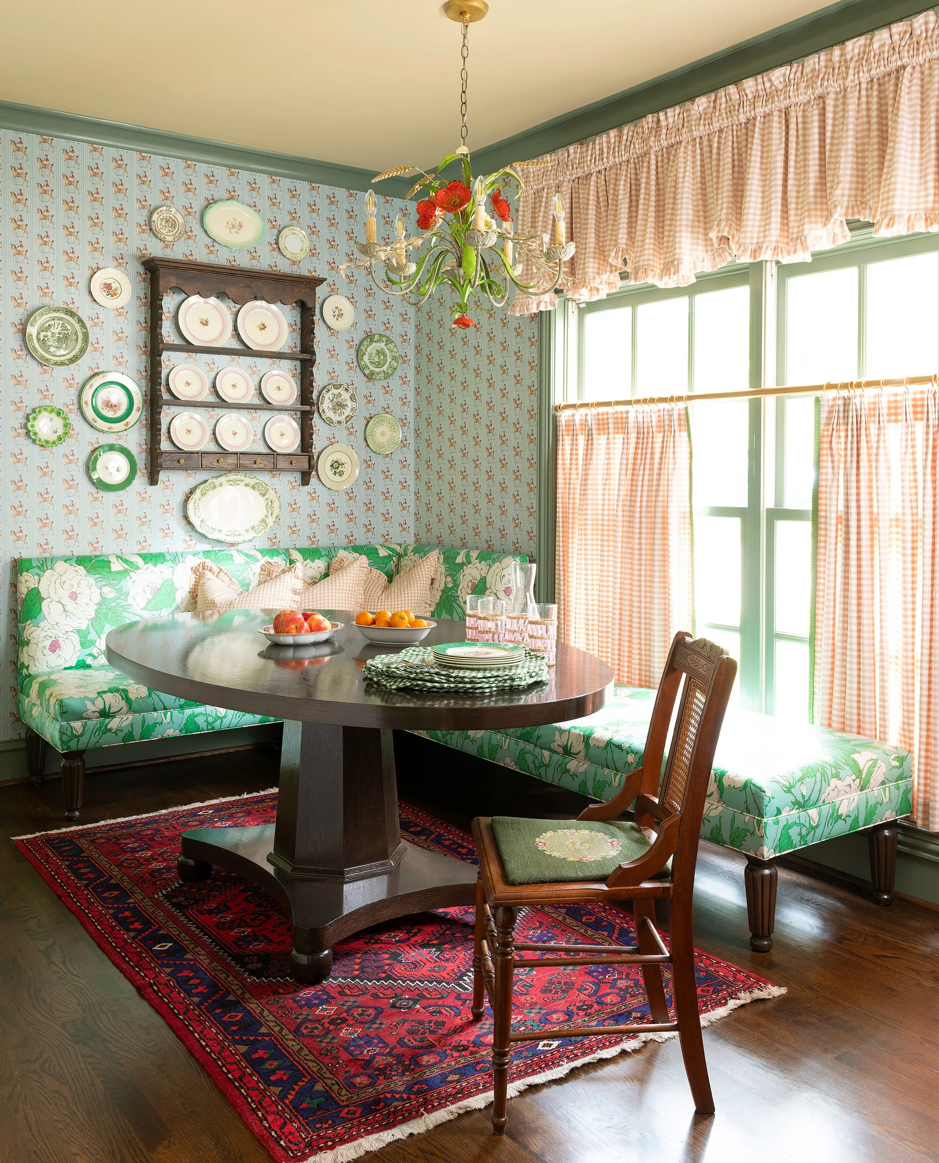 A traditional dining nook features a green floral upholstered banquette, an oval wooden table, pink gingham cafe curtains, and vintage wall plates.