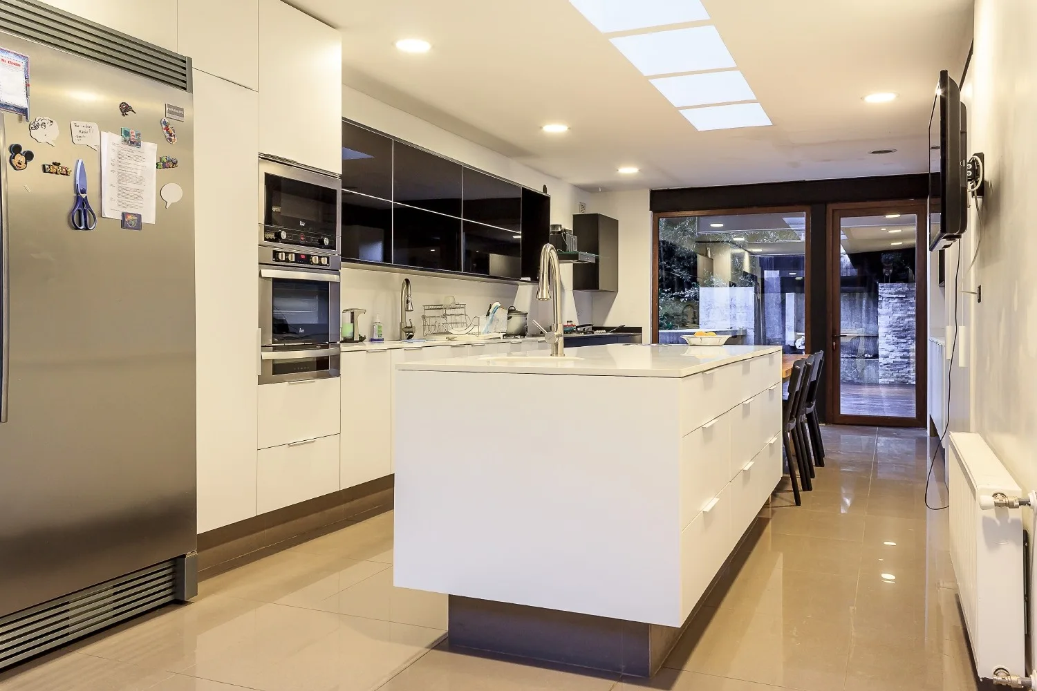 A modern kitchen featuring a large white central island, glossy black upper cabinets, stainless steel appliances, and ceiling skylights.