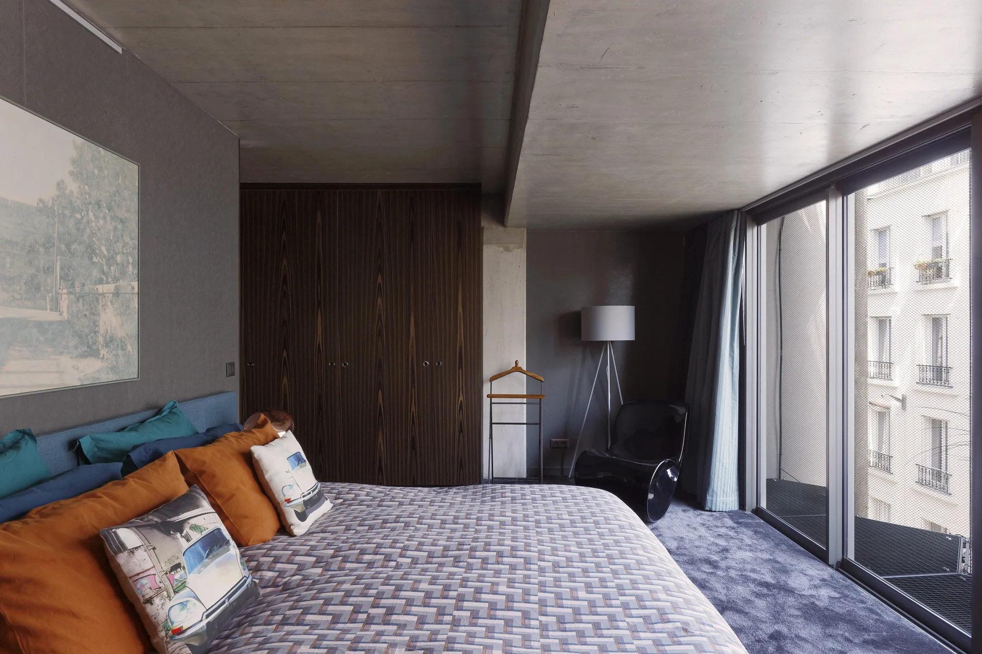 A modern bedroom features a bed with orange pillows, a dark wood built-in wardrobe, a black accent chair, and an exposed concrete ceiling.