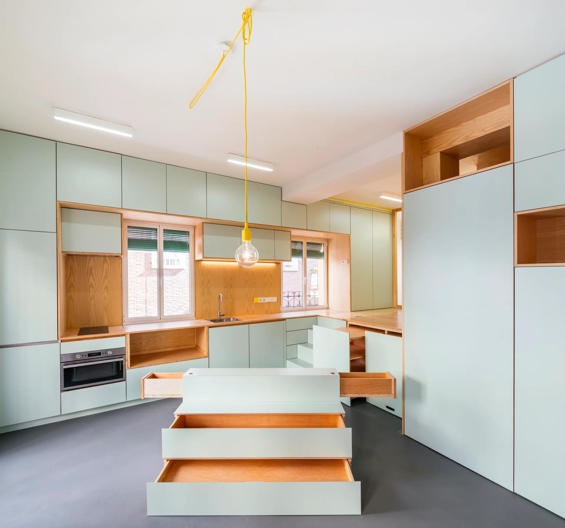 A minimalist kitchen features matte mint green cabinetry, warm natural wood open shelving, and a central island with pulled-out drawers.