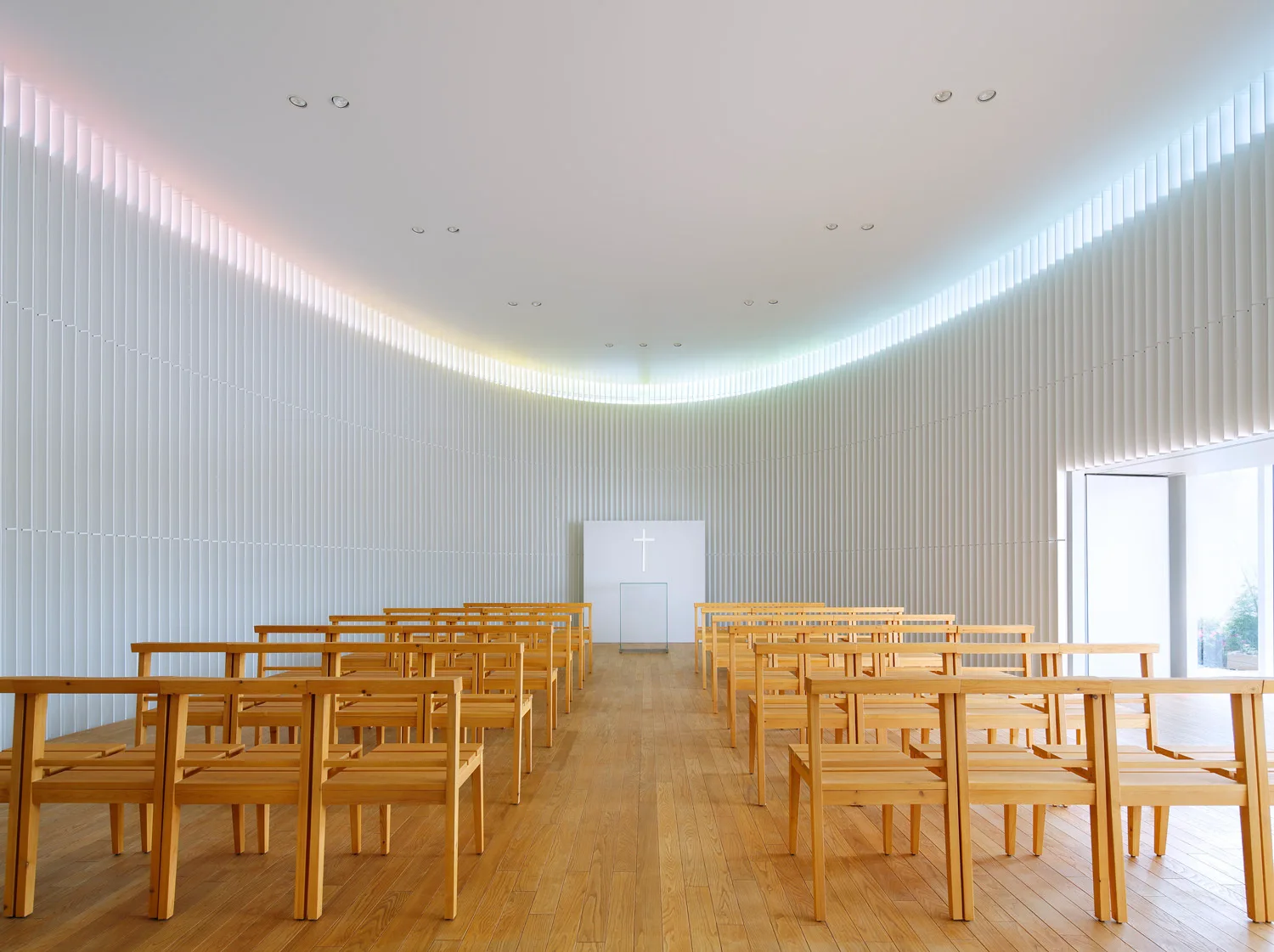 A minimalist chapel interior features rows of light wood chairs facing a simple white altar against a towering curved wall of white vertical slats.