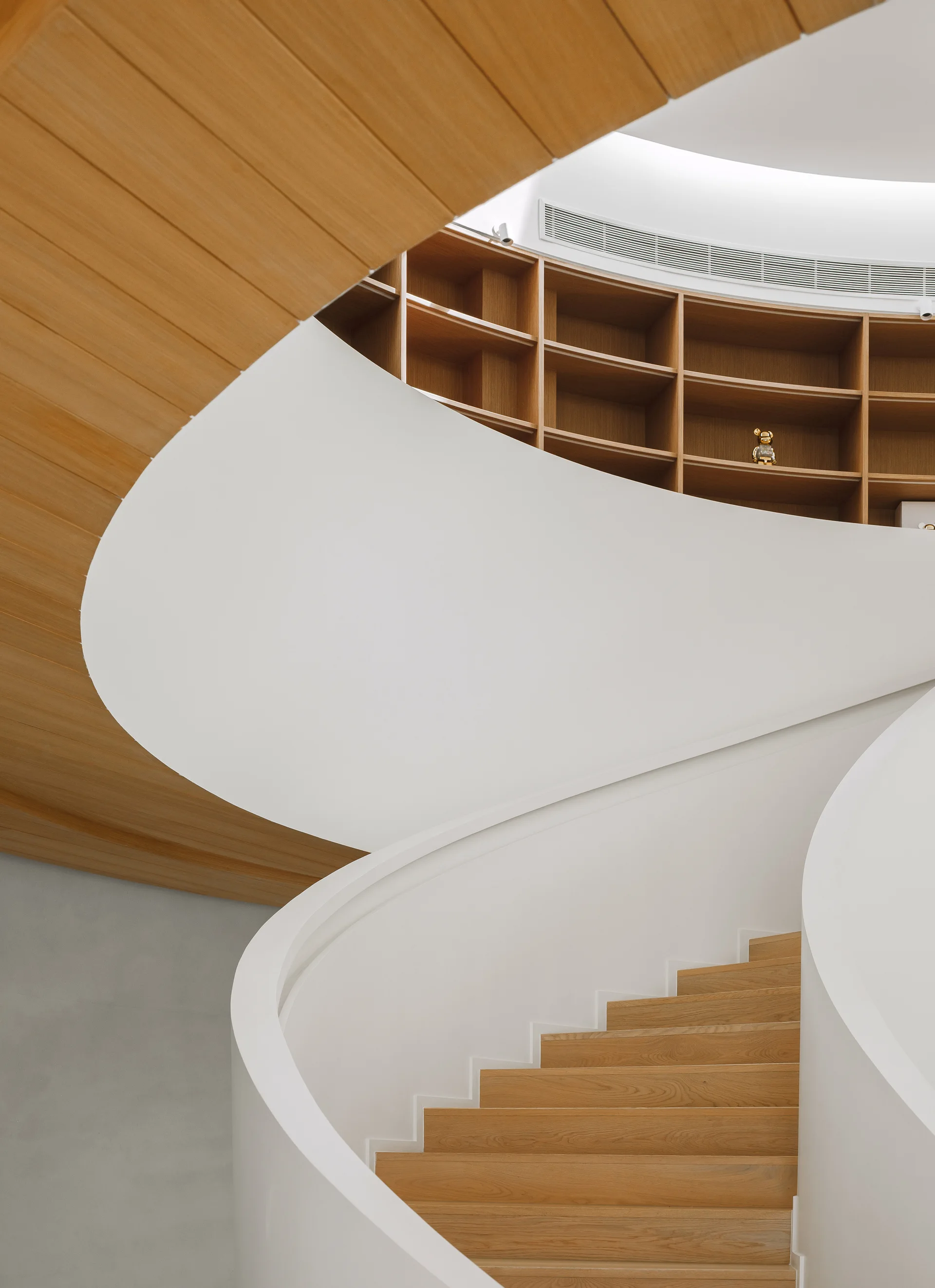 A minimalist spiral staircase with white railings and wooden treads winds up toward curved wooden shelving integrated into the wall.