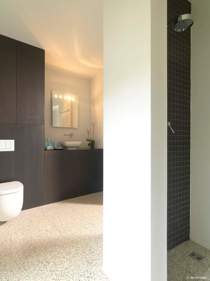 A minimalist bathroom featuring a dark wood-paneled vanity with a vessel sink, a wall-mounted toilet, and a walk-in shower with dark mosaic tile.