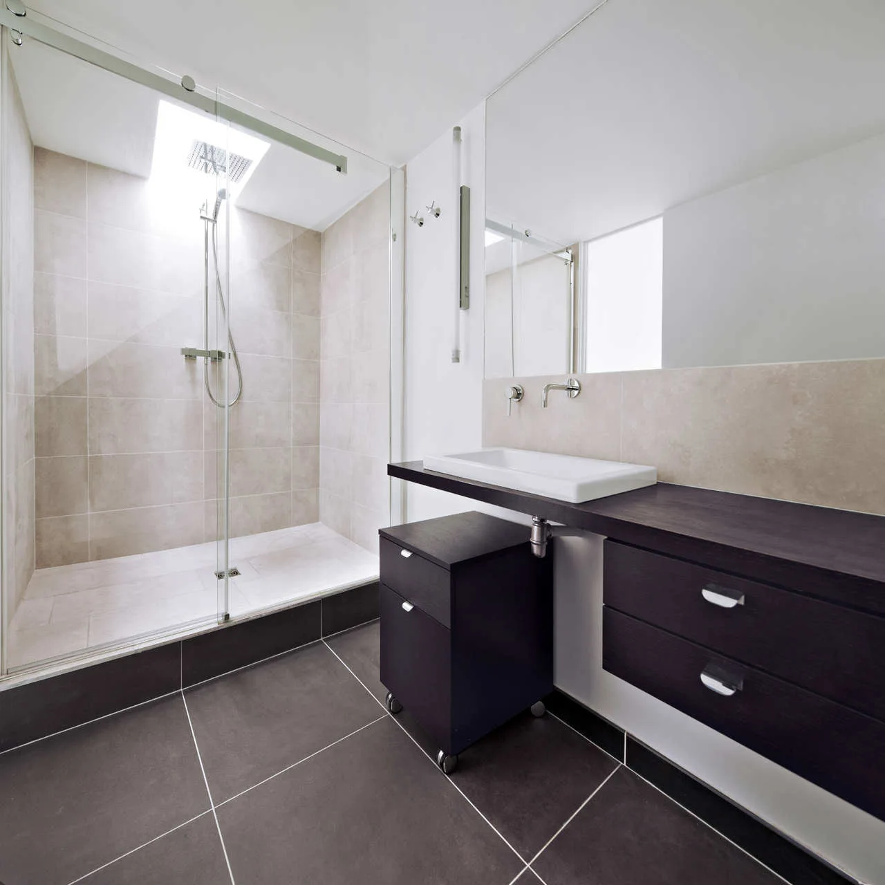 A modern bathroom with a glass walk-in shower under a skylight, a floating dark wood vanity with a white rectangular basin, and dark grey floor tiles.