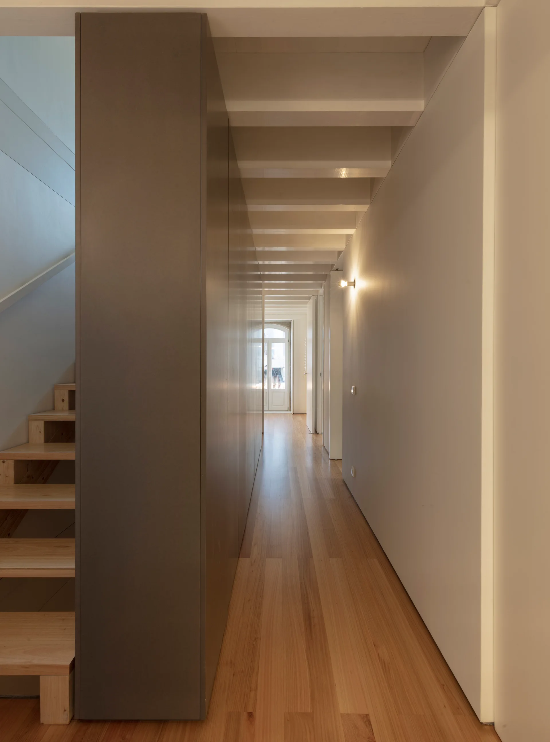 A minimalist hallway features a long grey built-in cabinet wall, exposed ceiling joists, and a light wood staircase on the left.